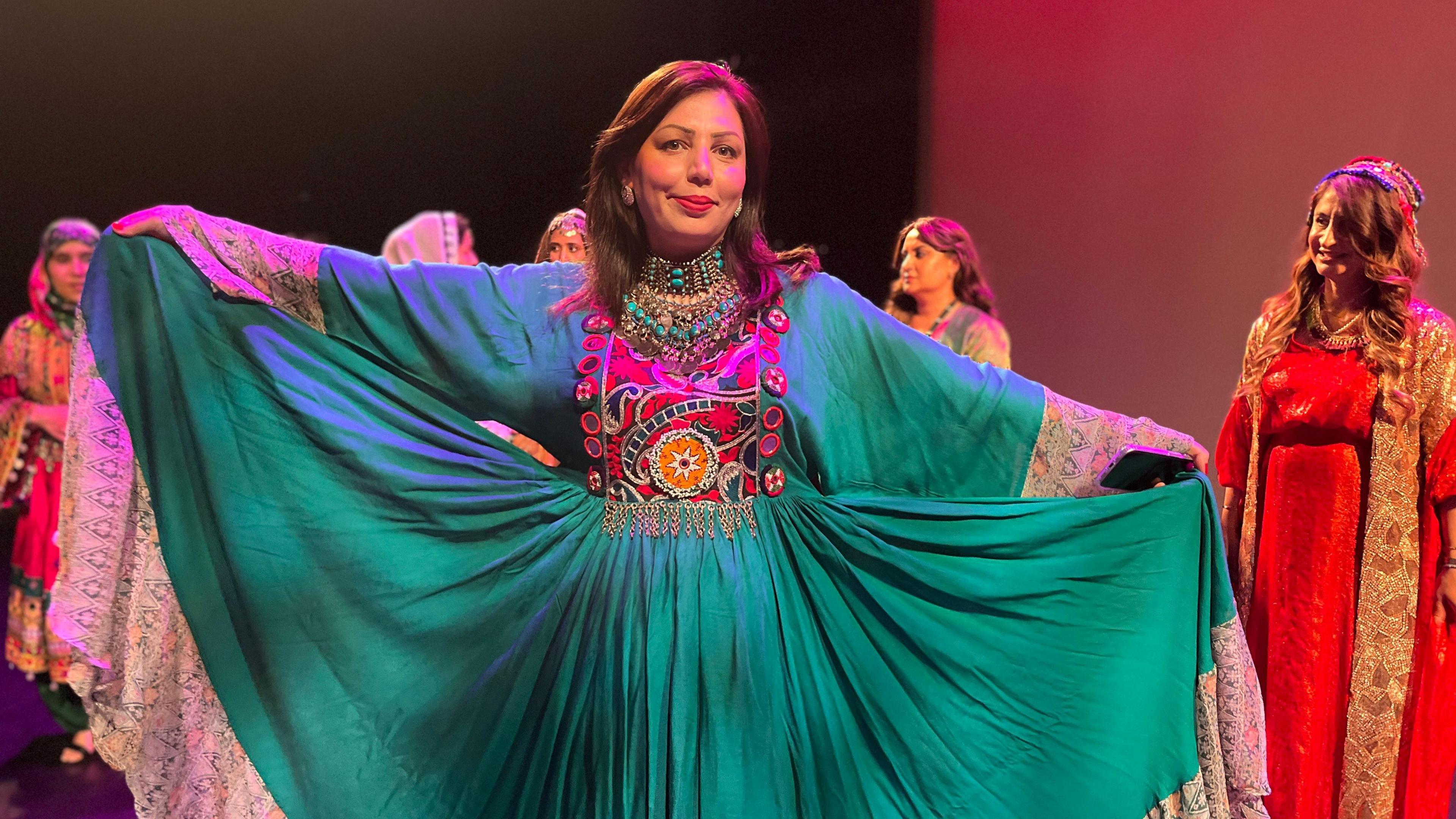 A woman wears a flowing green dress with colourful embroidery