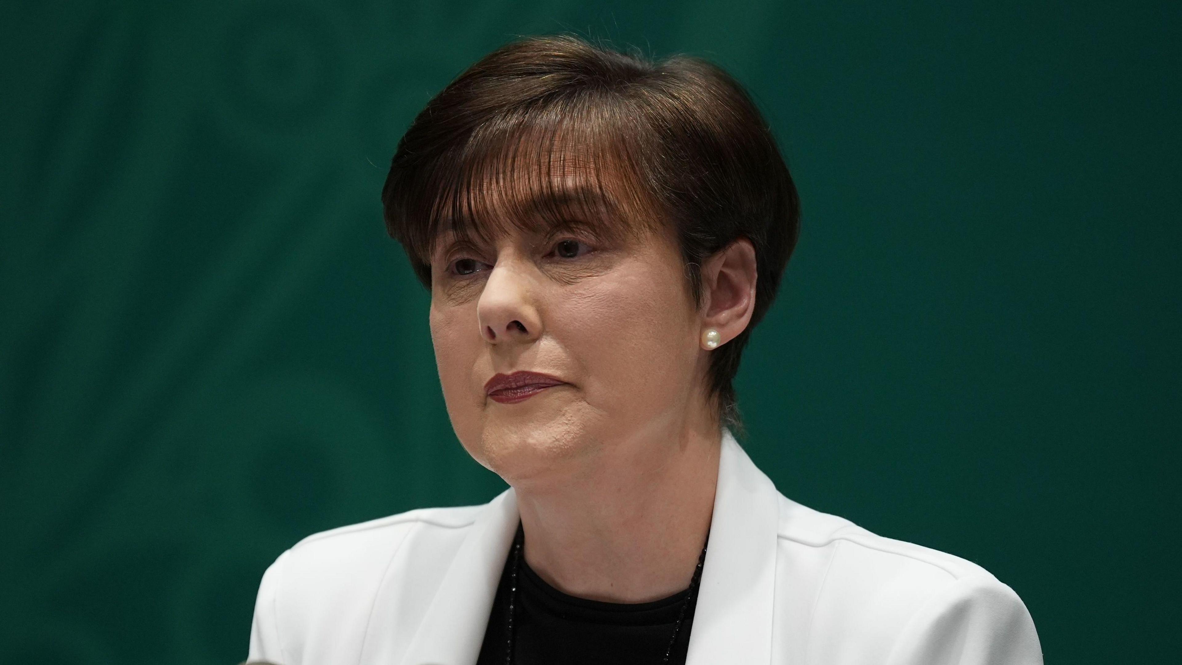 A woman with short brown hair wears a white blazer and black top as she speaks into two black microphones. Behind her is a green background. She wears white pearl earrings.