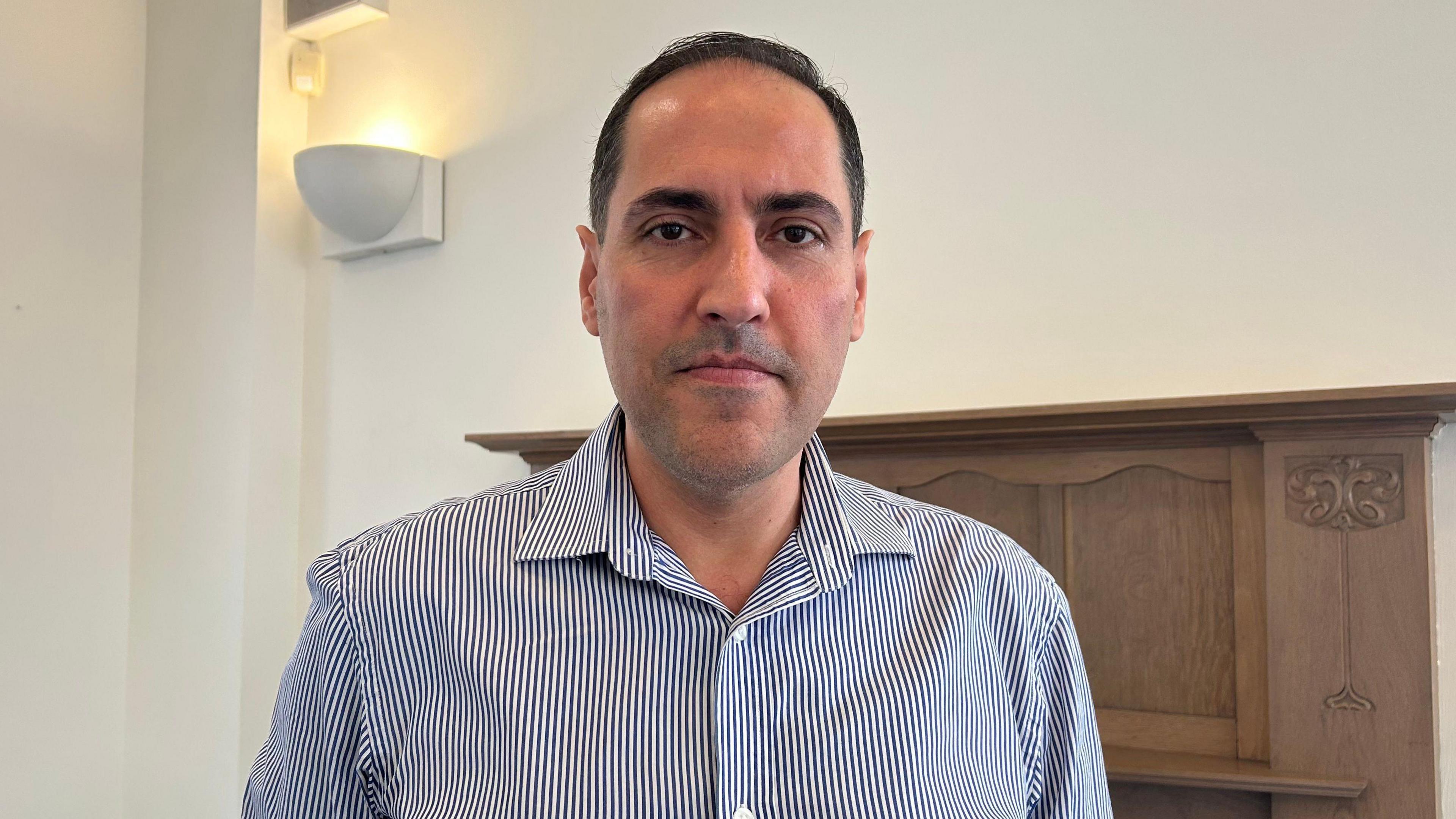 Dr Ahmed Sabra in a striped blue shirt. He has short black hair and is stood in front of a wooden mantle place. There is a light fixture to the left of him.