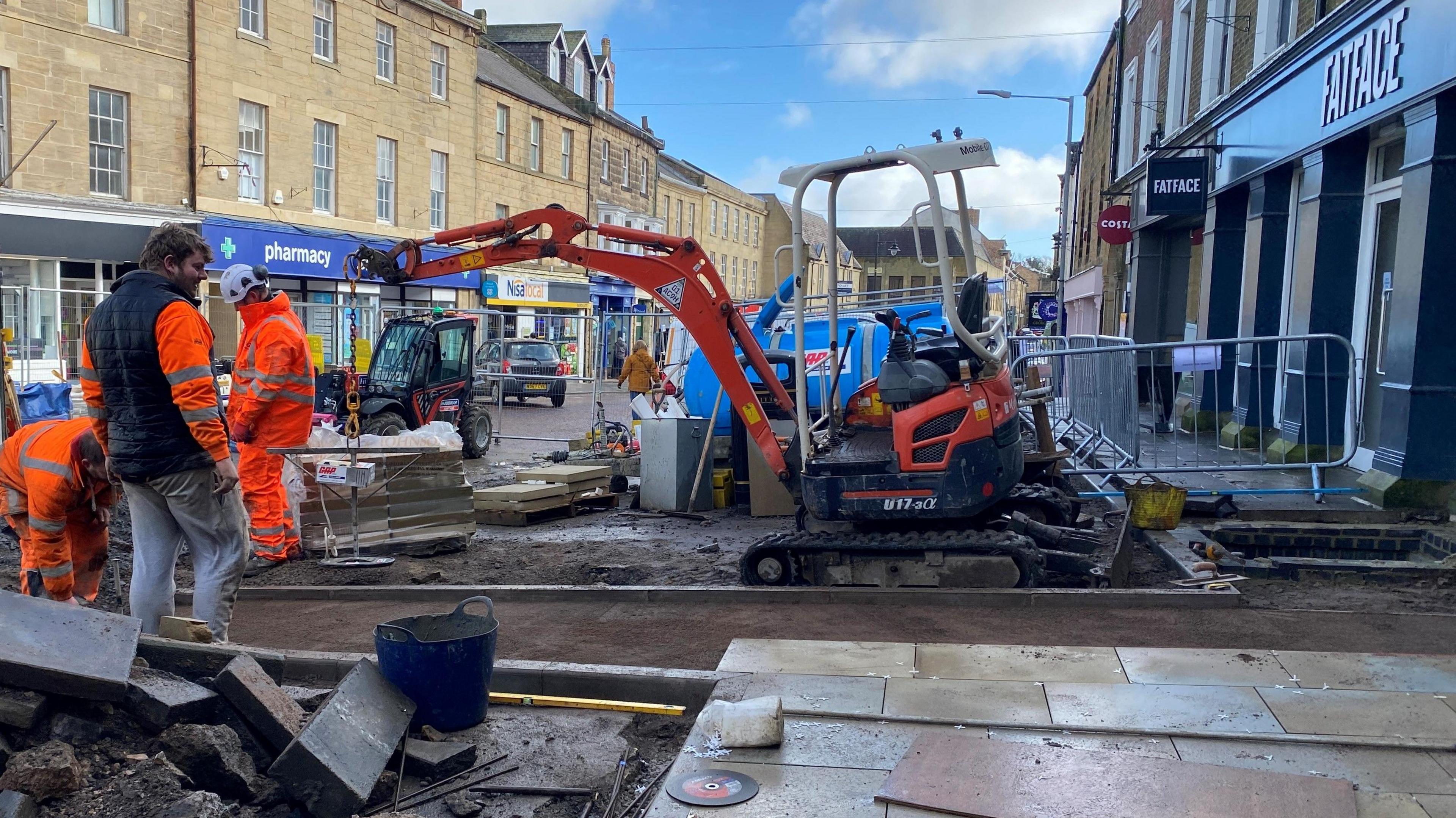 Workmen are working on a road with a digger and lots of paving stones. Three men are working surrounded by metal fences, they are all wearing hi-vis jackets  