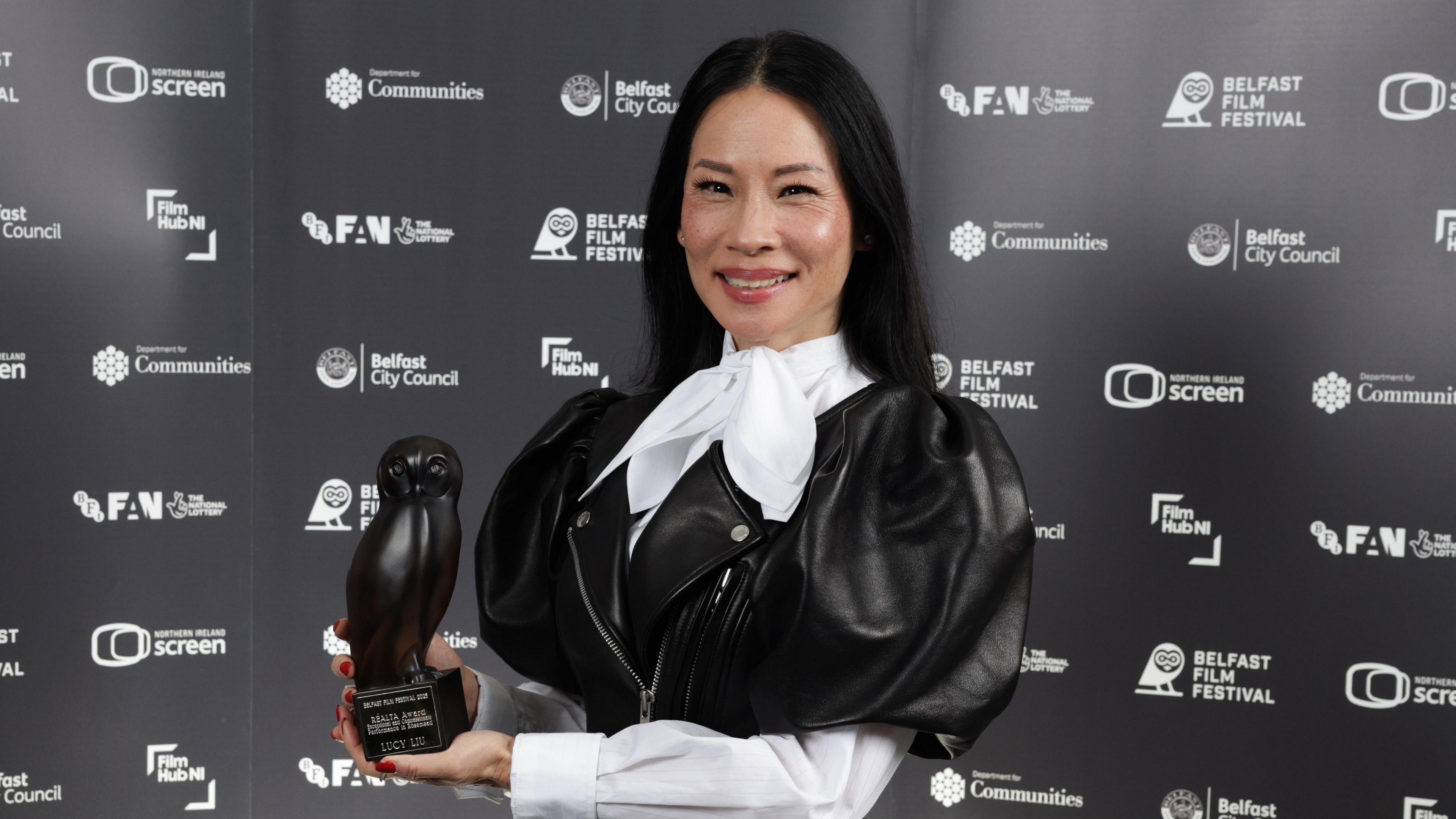 Lucy Liu smiles while posing with her award at the Belfast Film Festival.  She has long, straight, black hair and is wearing a black leather frock coat with puffed sleeves over a white blouse tied with a large bow at her neck.  She is holding an award shaped like an owl. 