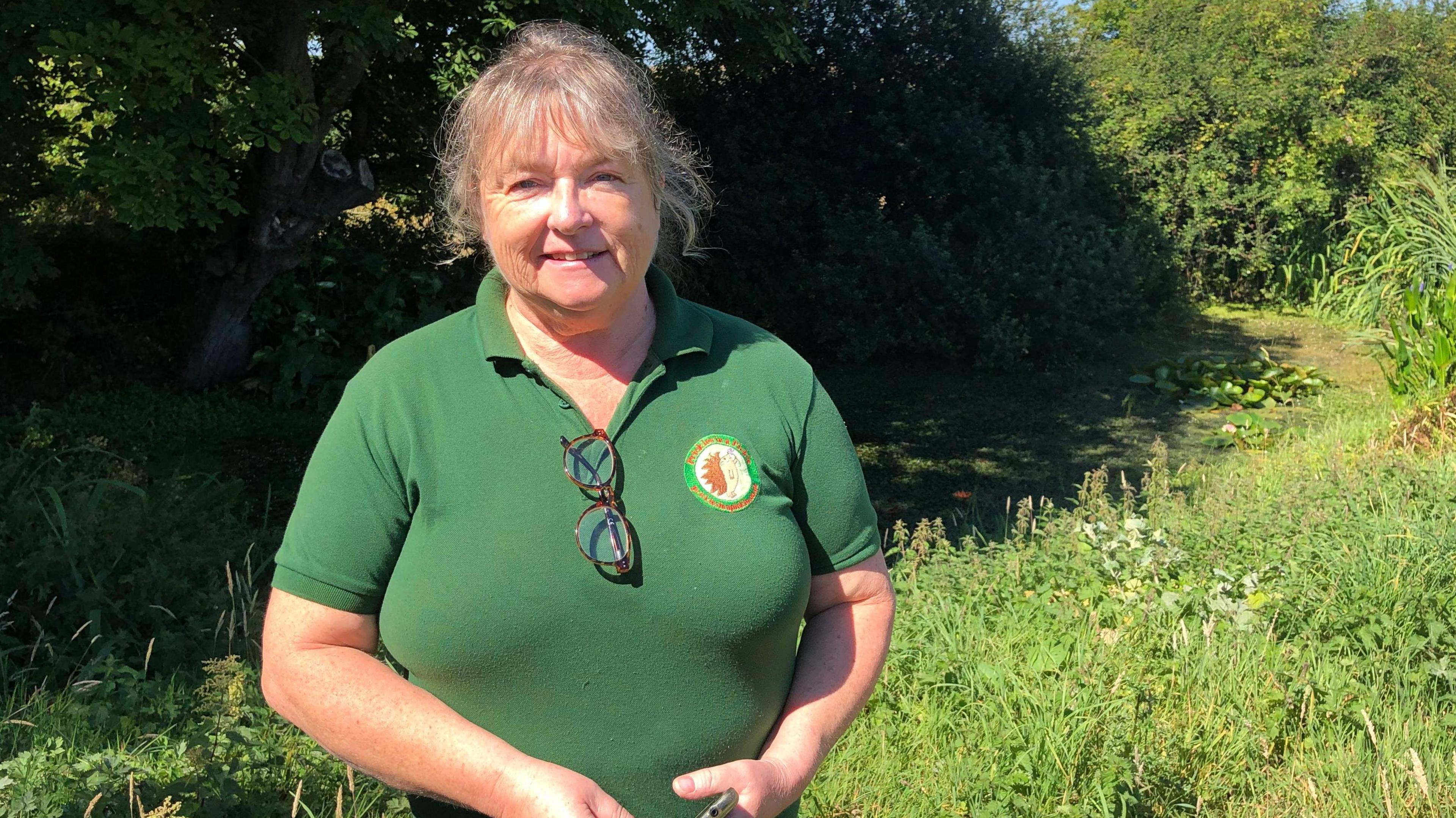 A picture of Judy in a field. She is wearing a green shirt with a logo on it. She has glasses hanging off her shirt and is smiling at the camera. Grass and trees can be seen in the background.