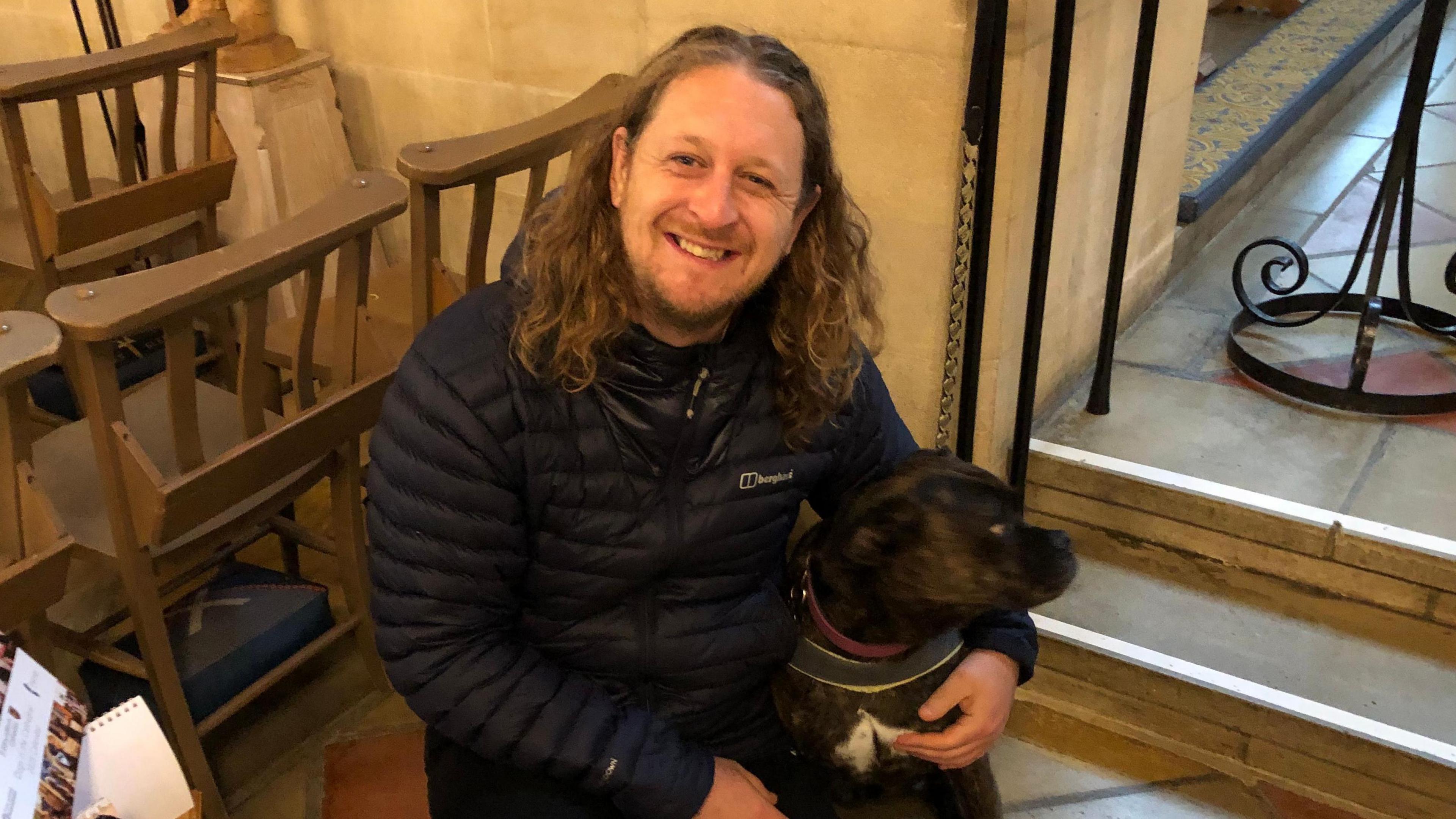 Adam Musgrove with shoulder length light brown hair and close cropped beard, smiling up at the camera, crouching down with an arm around his boxer/staffie cross who is looking away in St Edmundsbury Cathedral