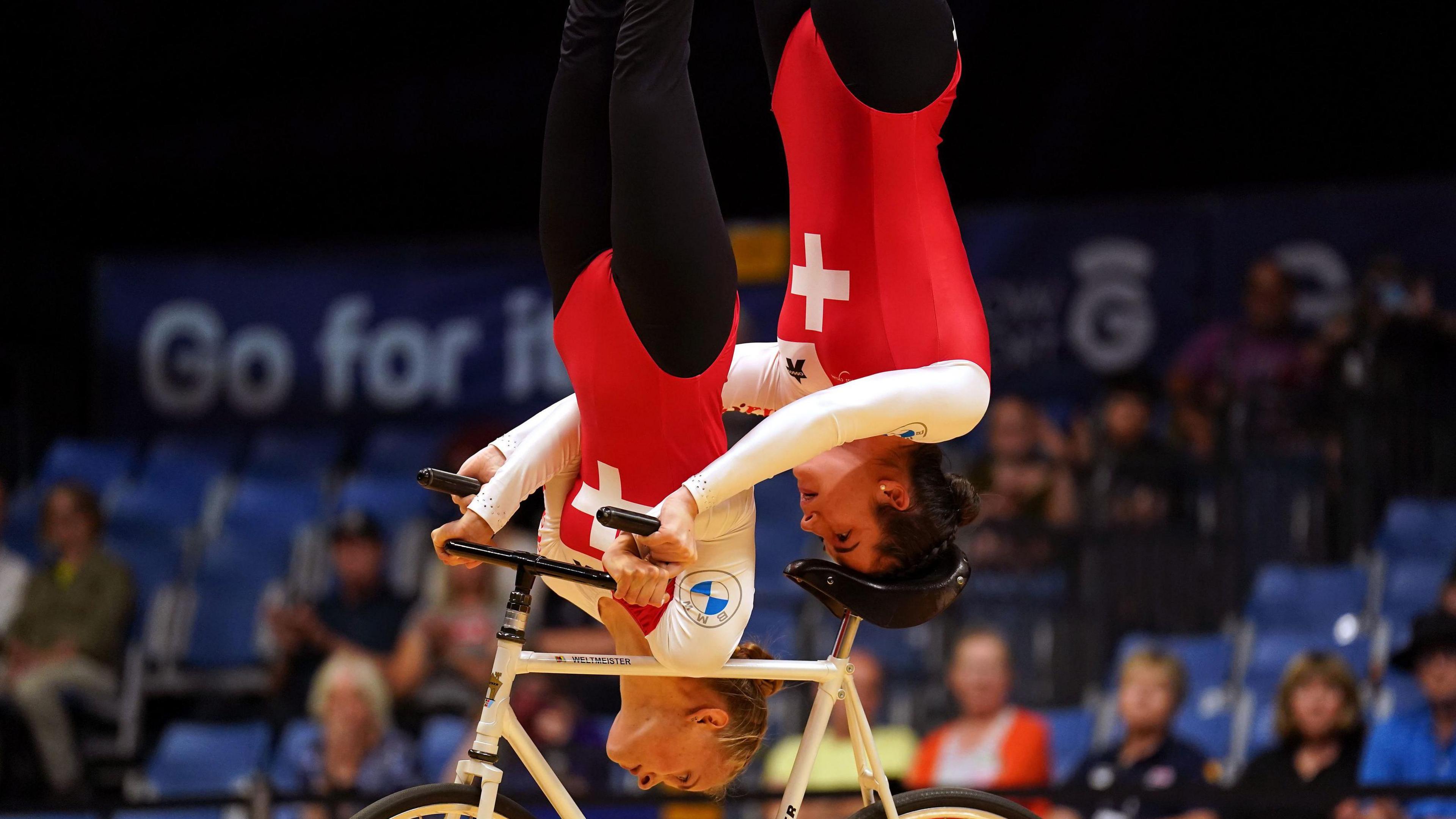 Two cyclists wearing Switzerland branded clothing perform an upside down trick on a bike with a crowd behind the cyclists