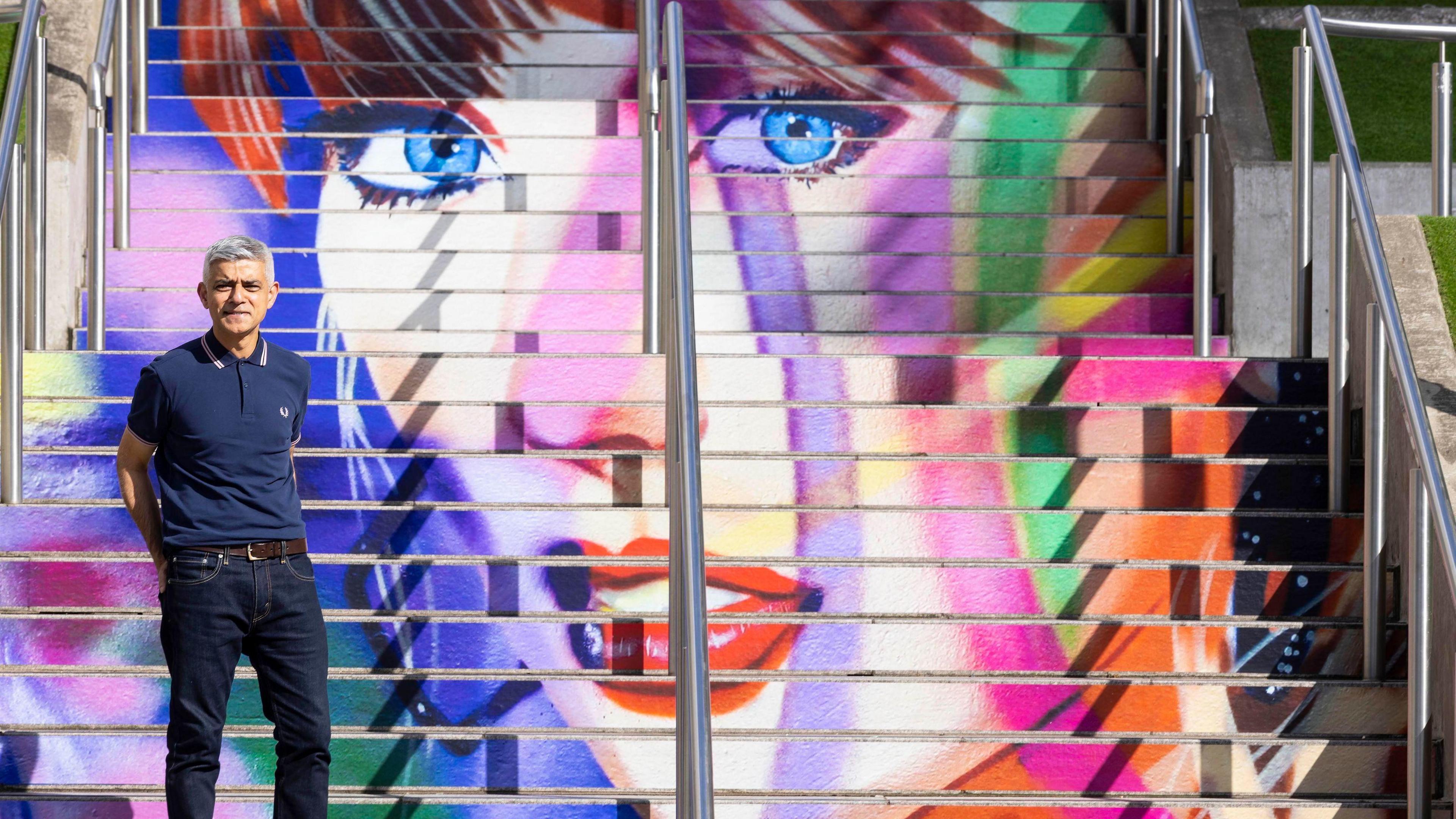 Sadiq Khan in a blue T-shirt standing on the steps of the multi-coloured Taylor Swift staircase art