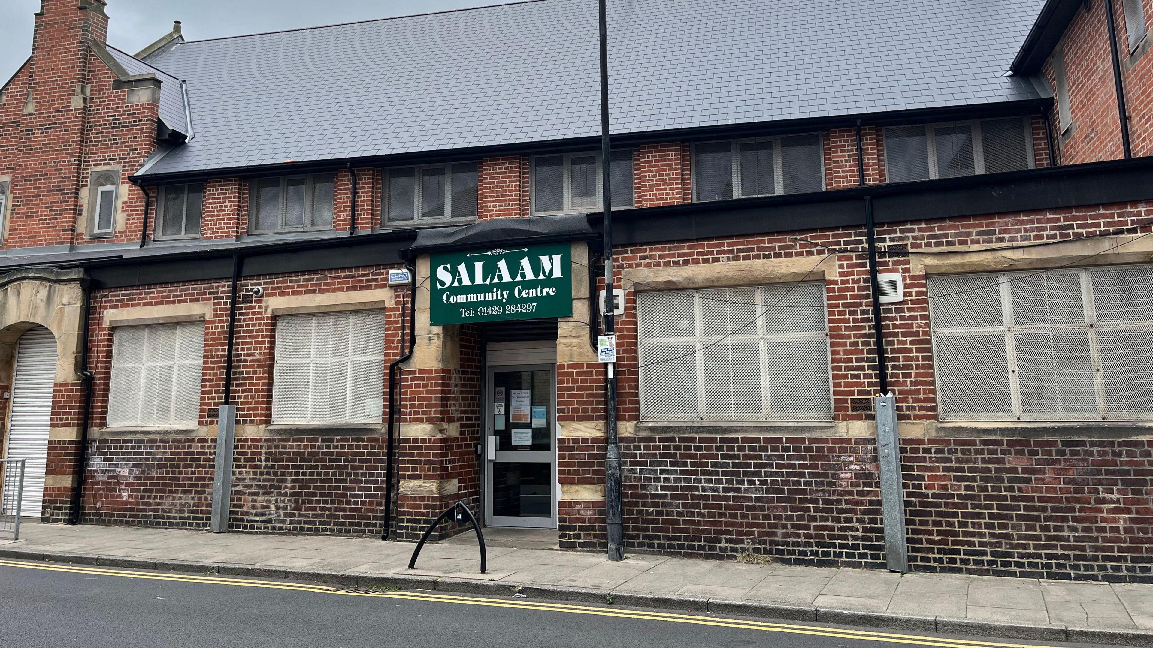 A red brick building on a road in Hartlepool which has a green Salaam Centre sign above the door. The windows are covered with white mesh.