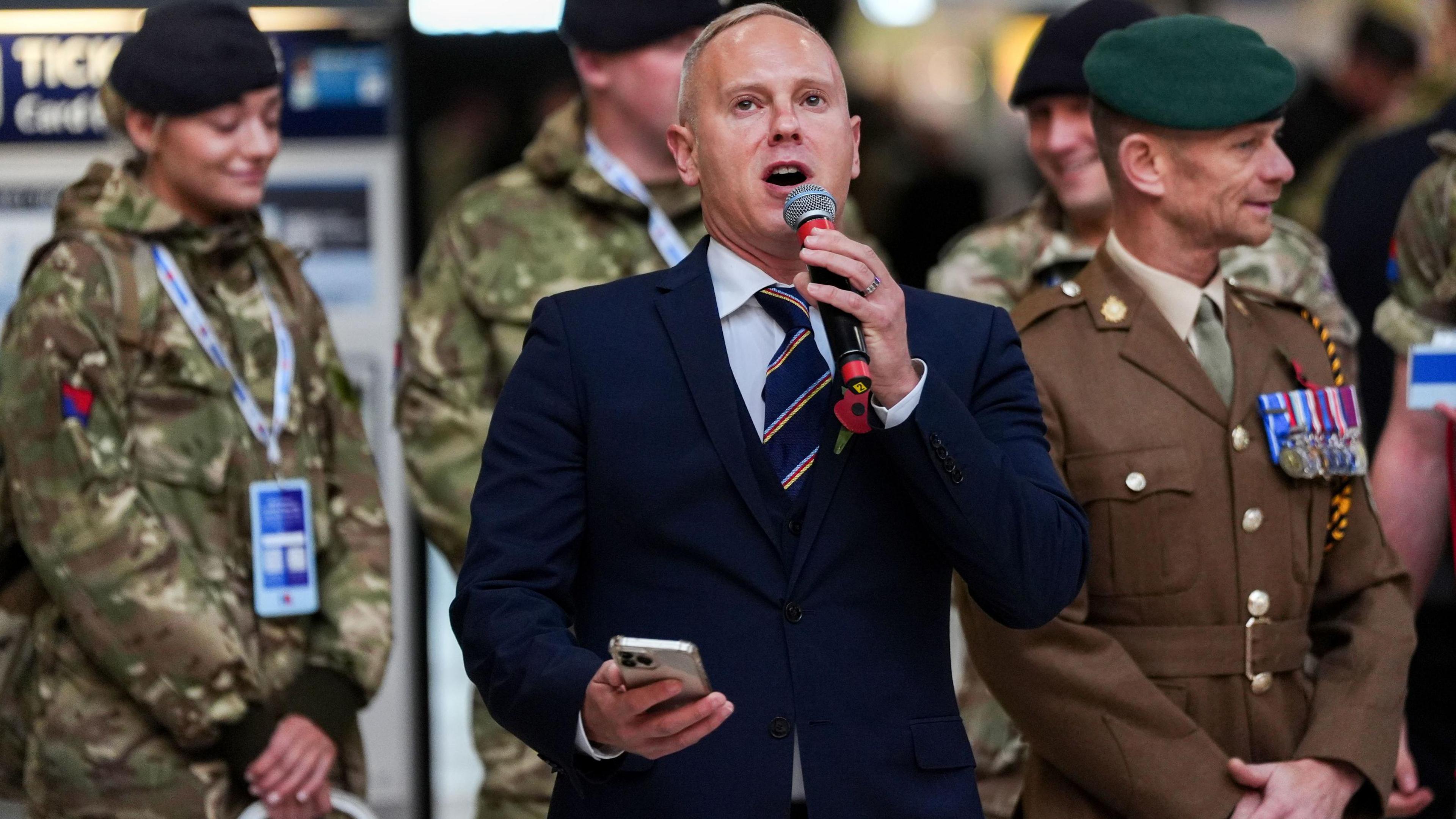 Dressed in a navy suit with a poppy in his lapel Rob Rinder holds a microphone and is talking to passengers. He is flanked by military personnel. A woman with a dark beret and a  green combat jacket is to the left of the picture with a man in a green beret and a brown military jacket stands to the right.