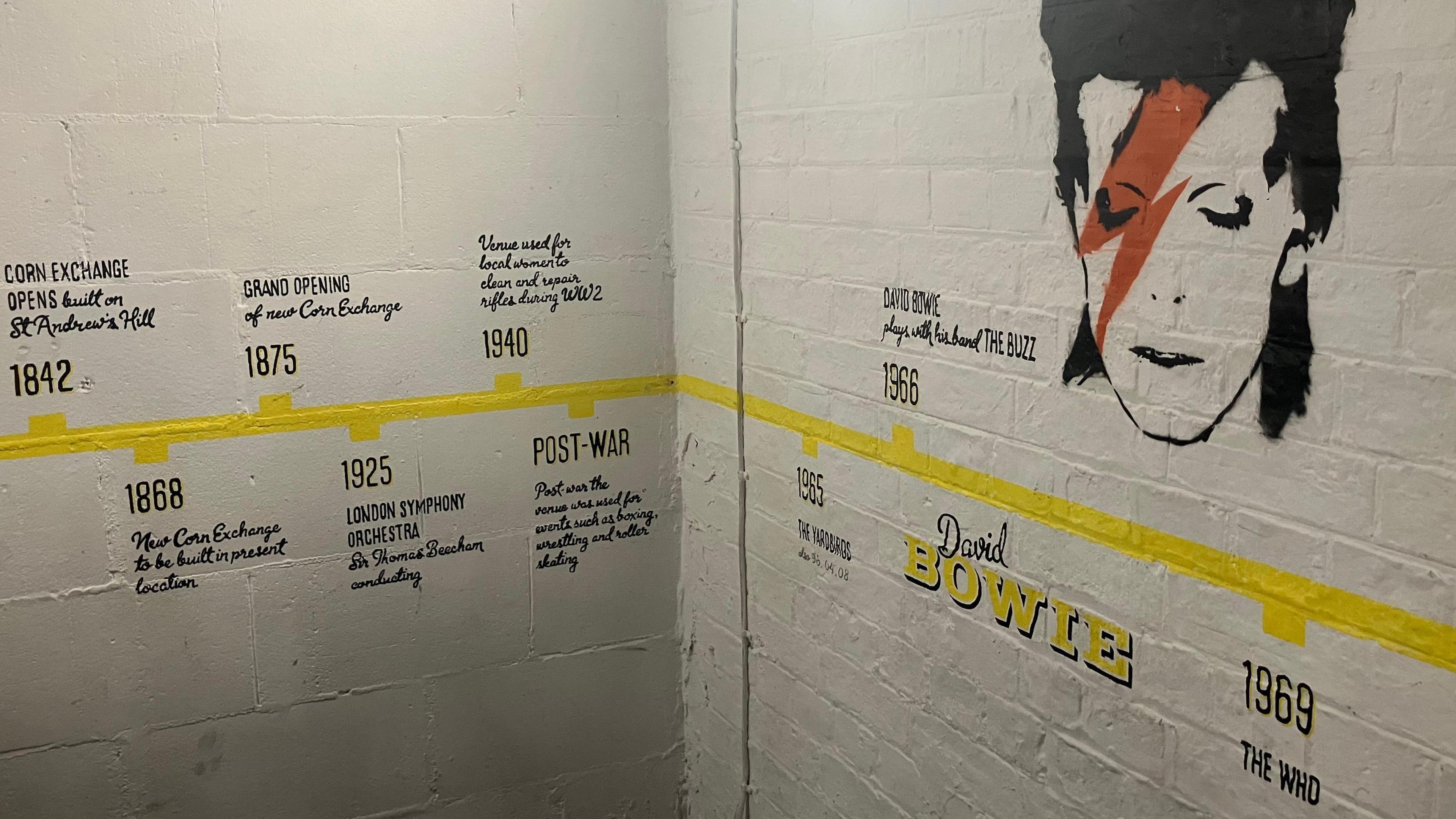 A bold yellow timeline stretches horizontally across a white bricked wall, marking key events from 1842 to 1969, including the venue’s grand opening, wartime dances, and performances by legendary artists like The Who. Each date is paired with black text detailing its significance. Dominating the right side of the mural is a striking illustration of David Bowie, featuring his iconic lightning bolt makeup, with his name boldly printed below.