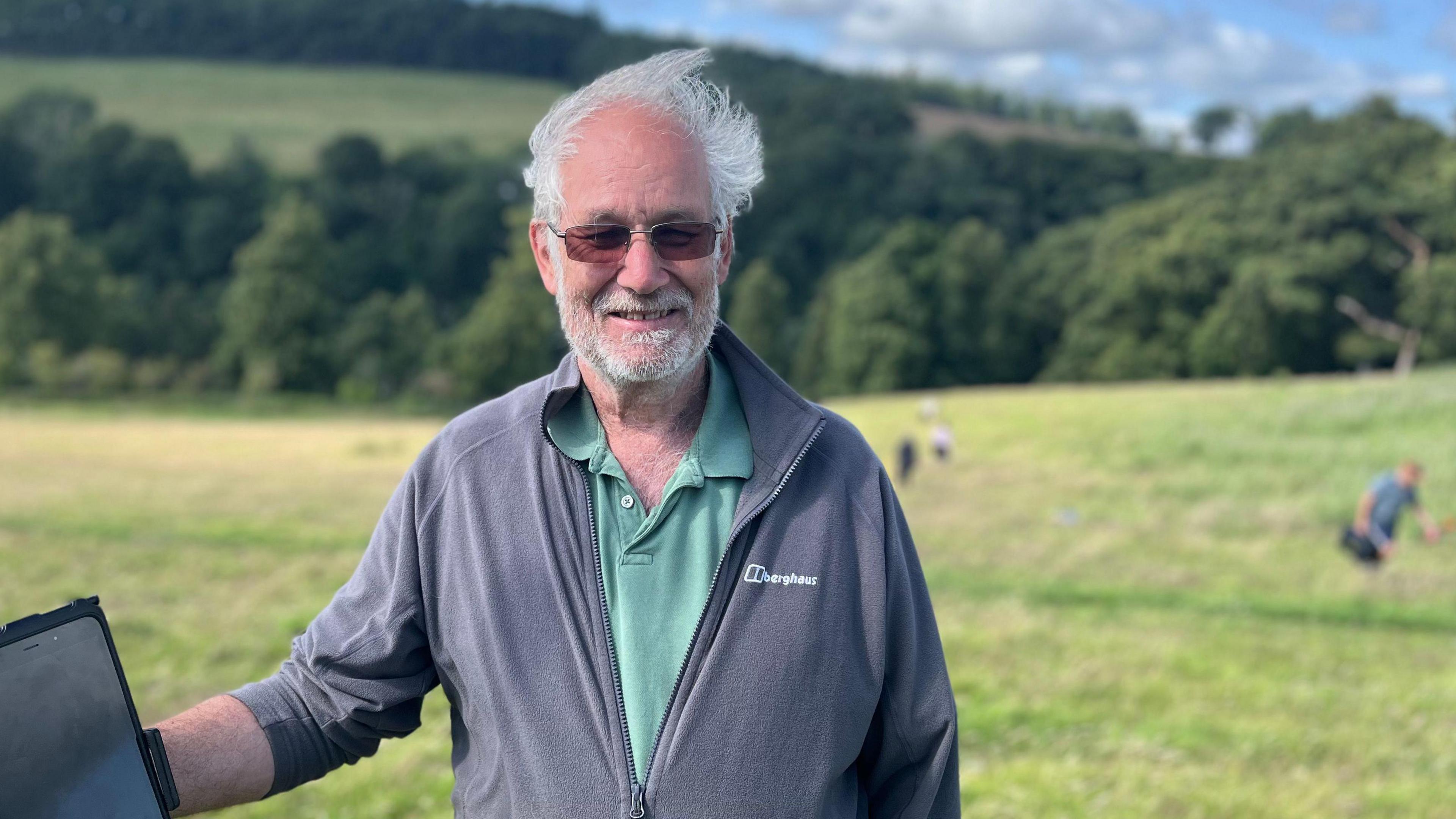 A grey haired man with dark glasses and a beard smiles at the camera. He's wearing a grey jacket with a green shirt beneath. Behind him are people working in a field.