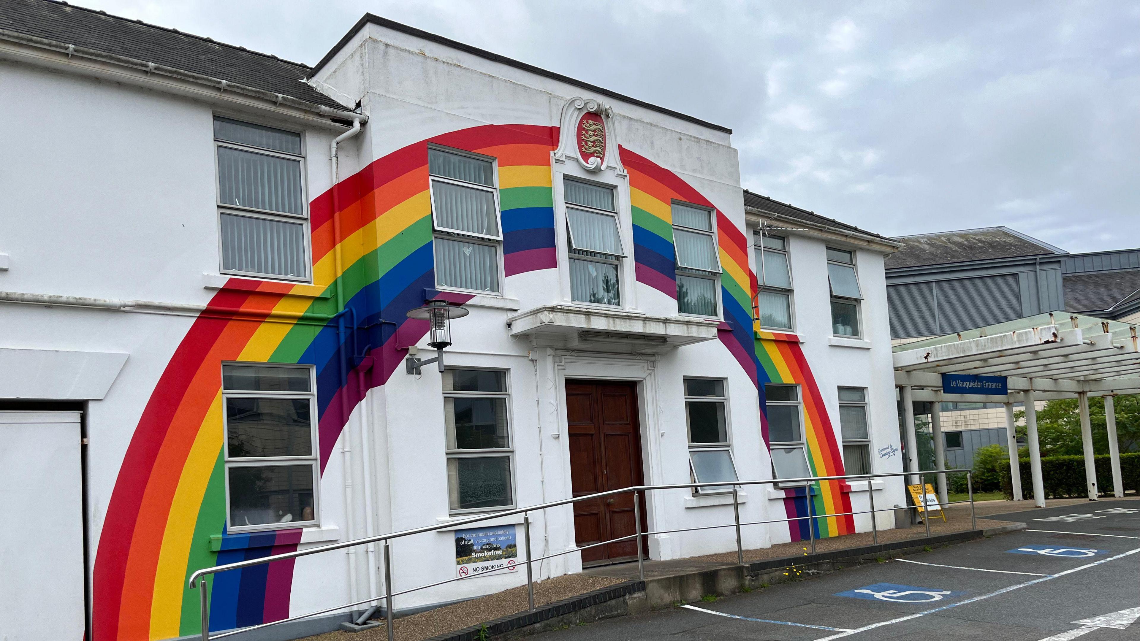 The entrance to Guernsey hospital. It shows the main entrance and a white builing to the sidem which is painted with a large rainbow flag. 