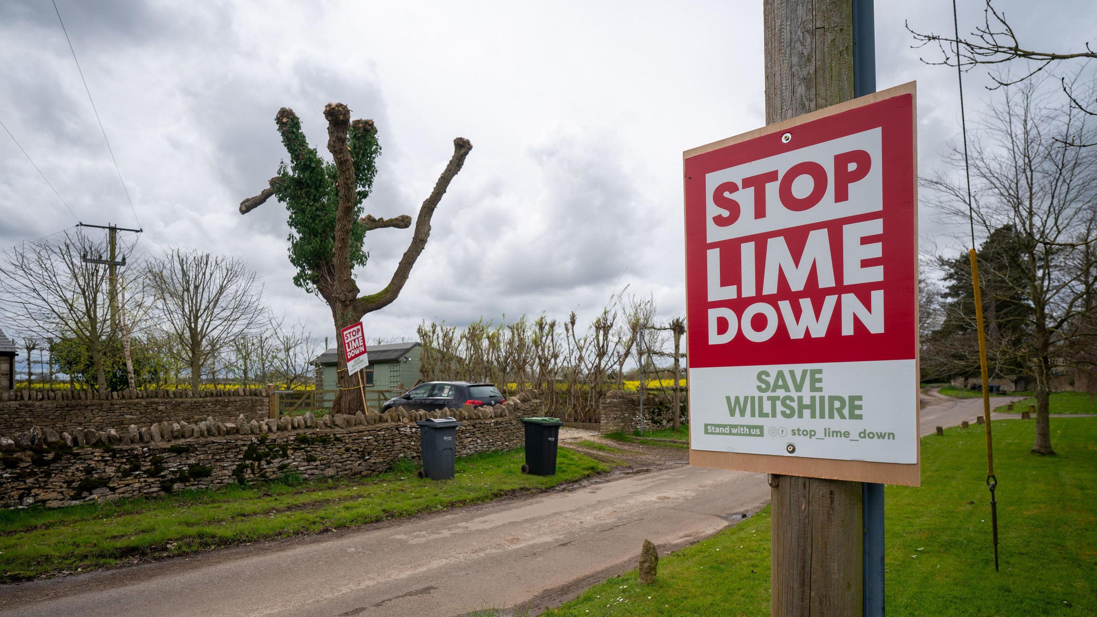 A sign in red and white which reads 'Stop Lime Down' and 'Save Wiltshire' attached to a post by a rural path.