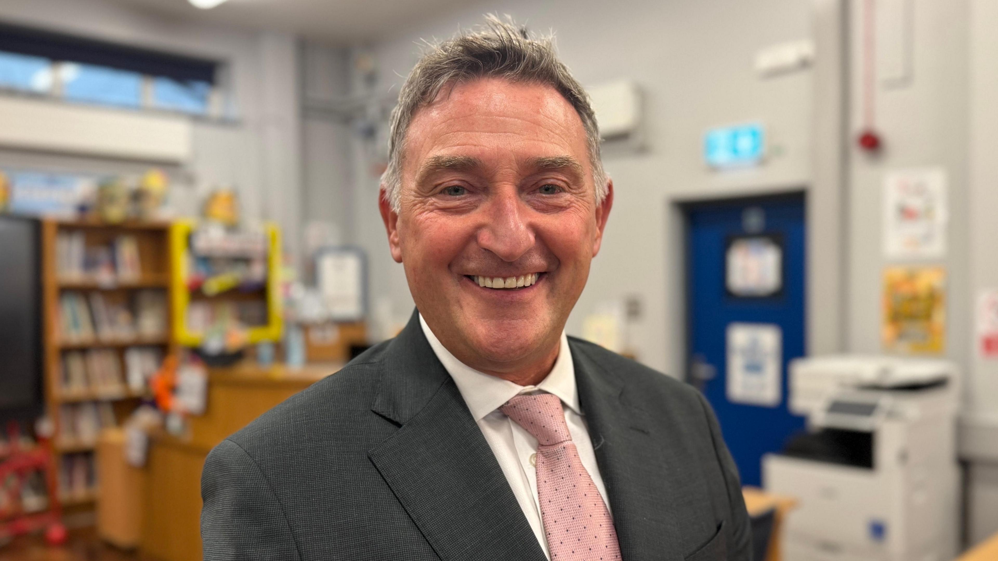 A man standing in a classroom smiles to the camera. He is wearing a grey suit, white shirt and pink tie. A book shelf and photocopier can be seen out of focus in the background as can a blue door with an emergency exit sign above.