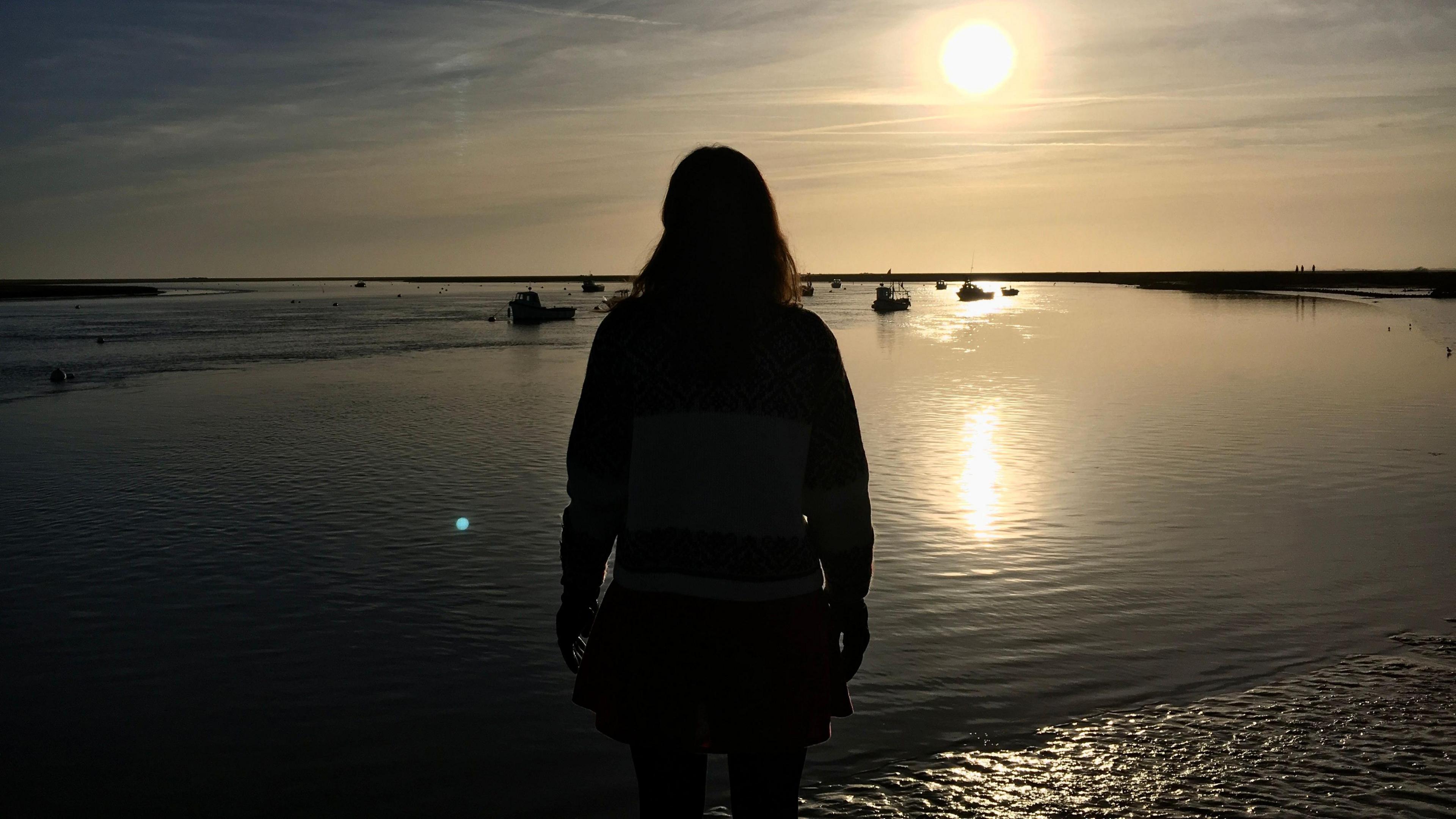 A woman stands on the quay at Orford in Suffolk, silhouetted against the sun. The water in the quay is at low tide and several boats are moored in the distance