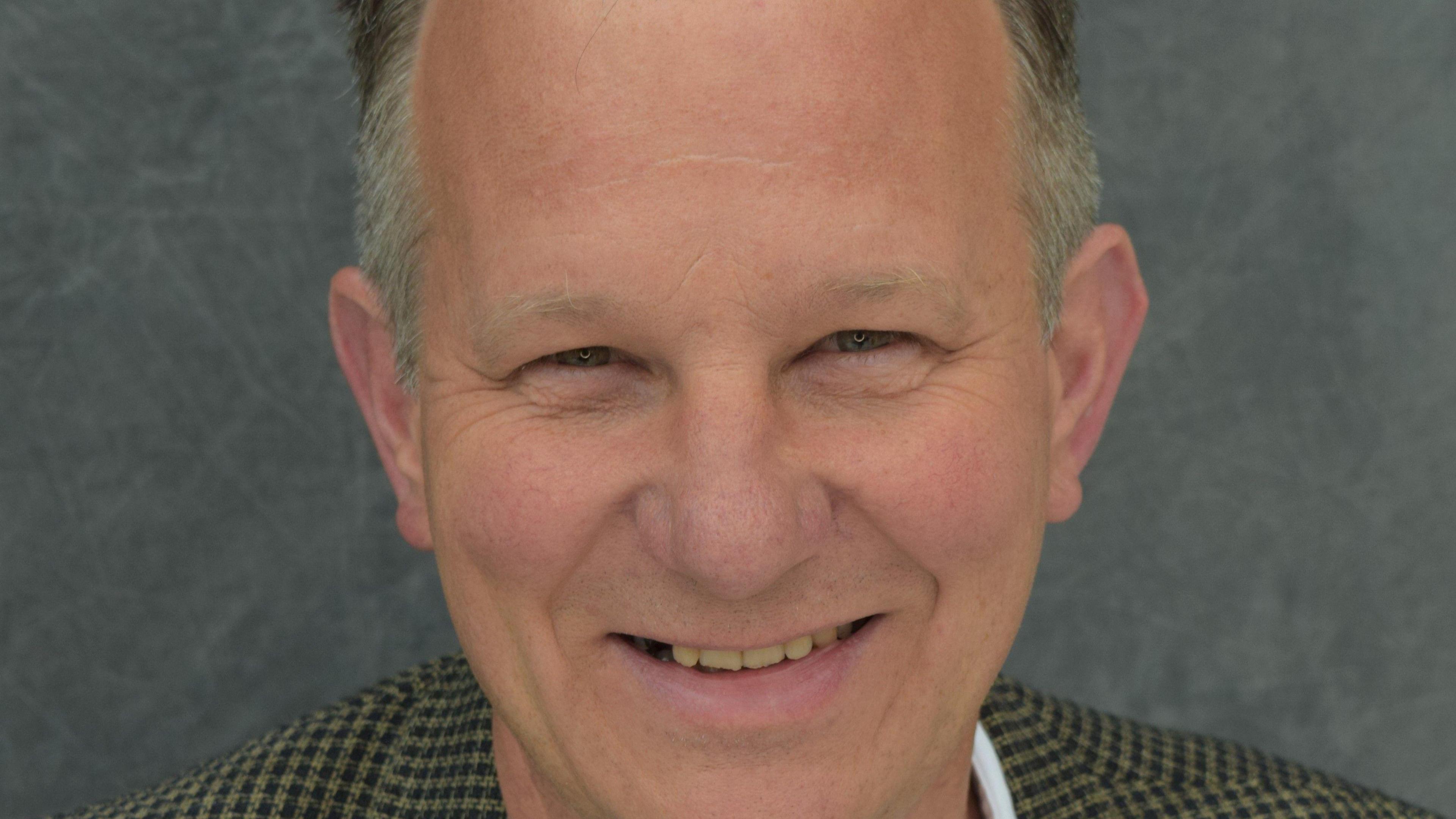 Close-up image of a man smiling. He is wearing a check jacket.