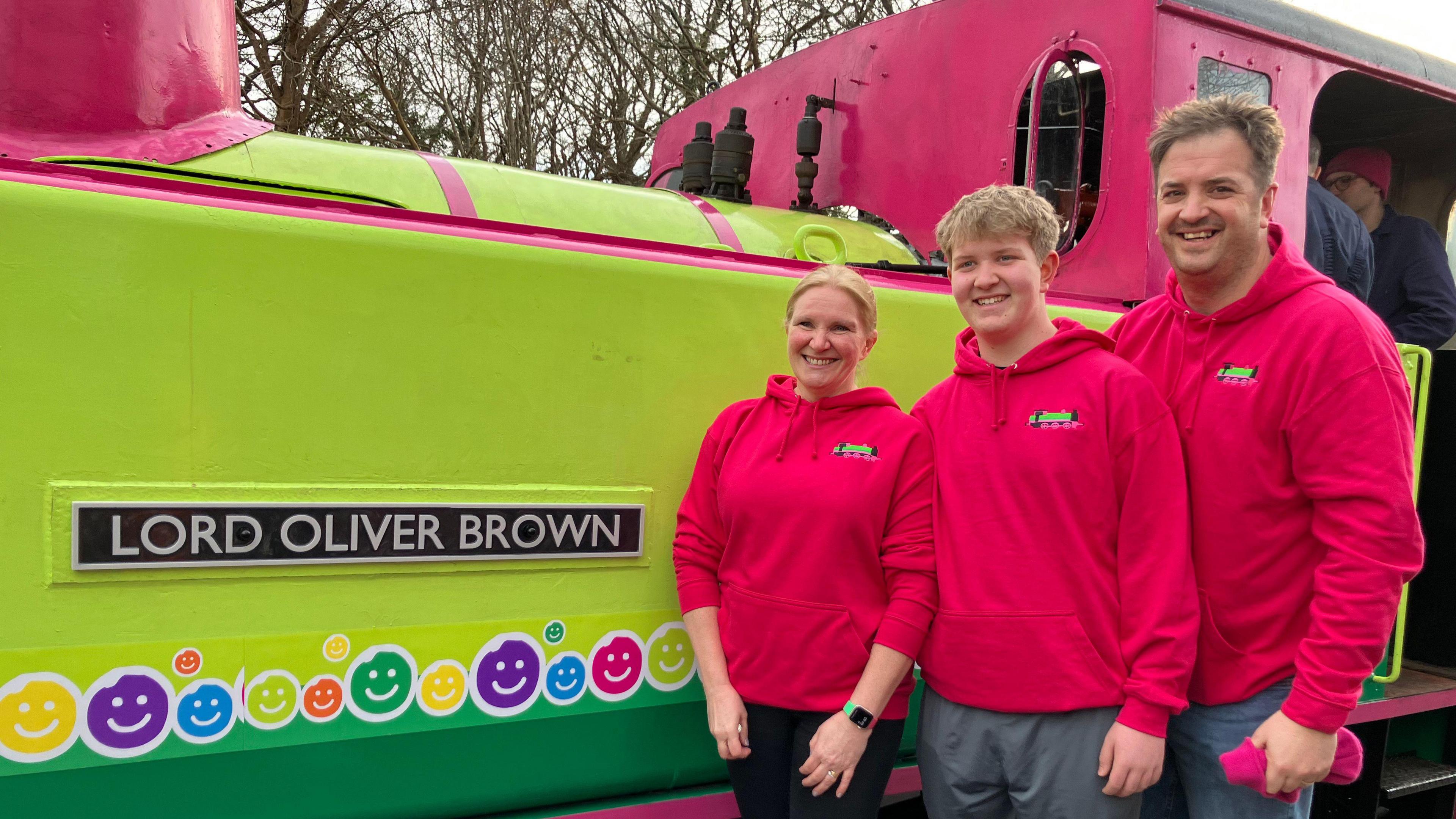 Nicky Brown, Ben Brown and Mike Brown pictured in front of the steam train. They are wearing bright pink hoodies with the green and pink train behind them.