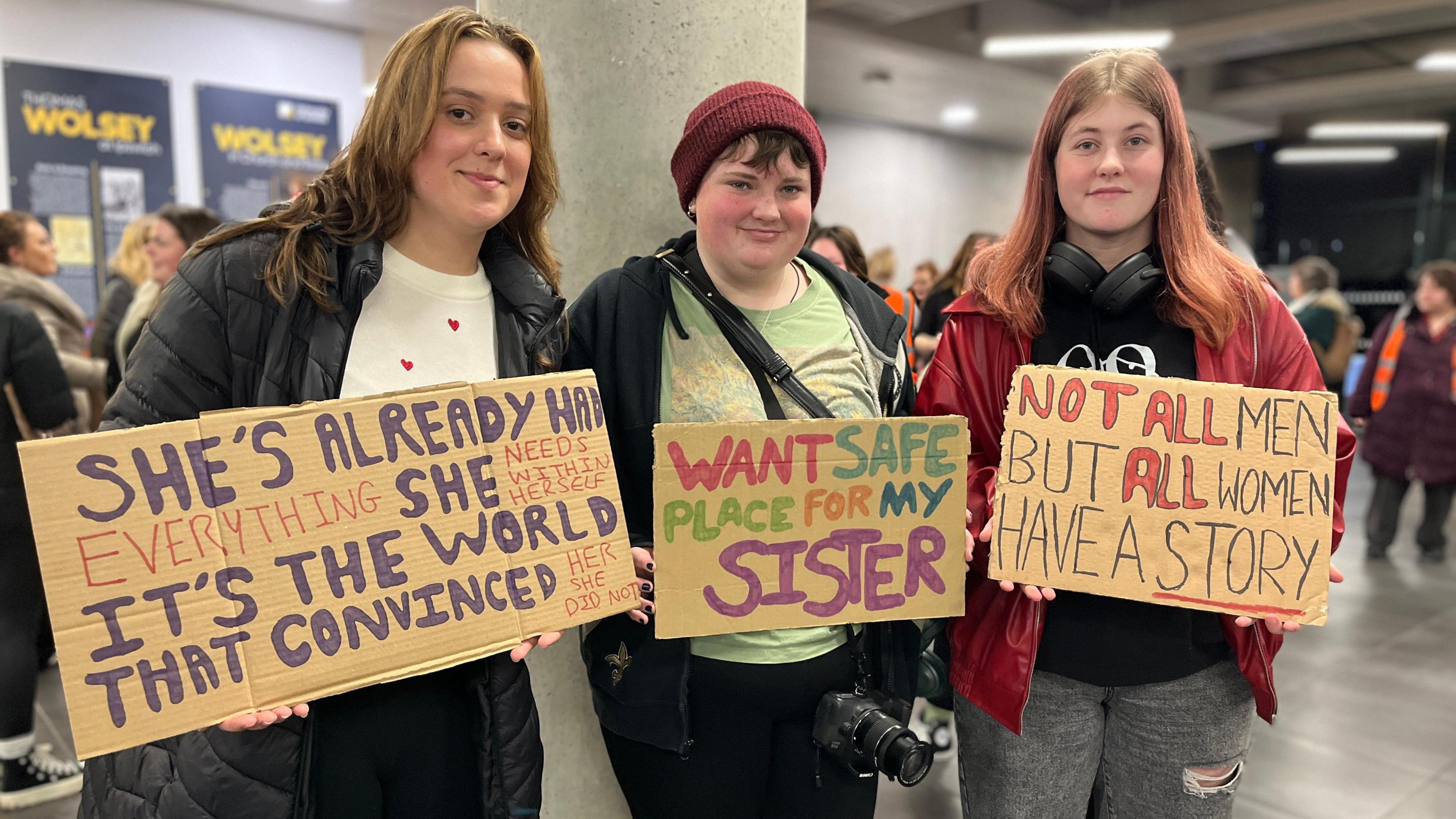 Three people hold cardboard banners with slogans written on them saying Not all men but all women have a story, want safe place for my sister and she's already had everything she needs within herself it's the world that convinced her she did not.