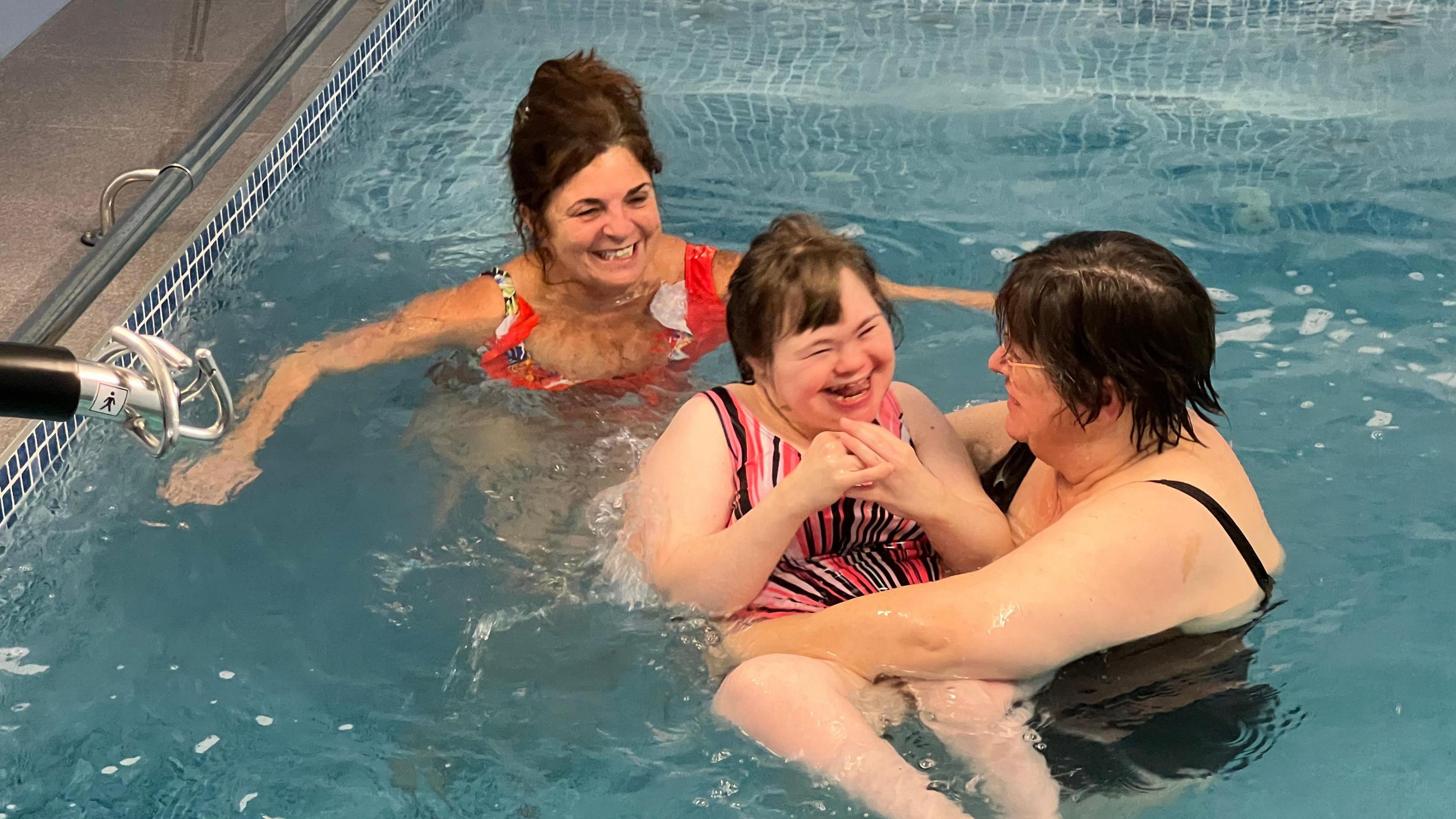 Three women are in a pool. One of the women is holding the woman in the centre, who is laughing. The woman behind them is smiling. 
