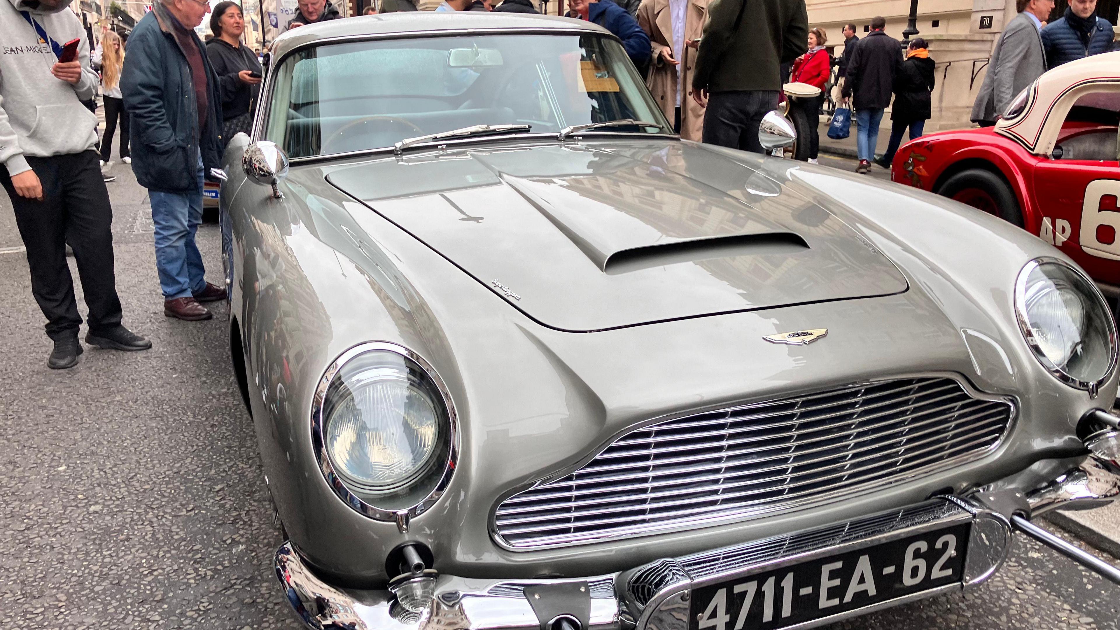 A silver Aston Martin DB5 is parked on pall mall as spectators, old and young, look on