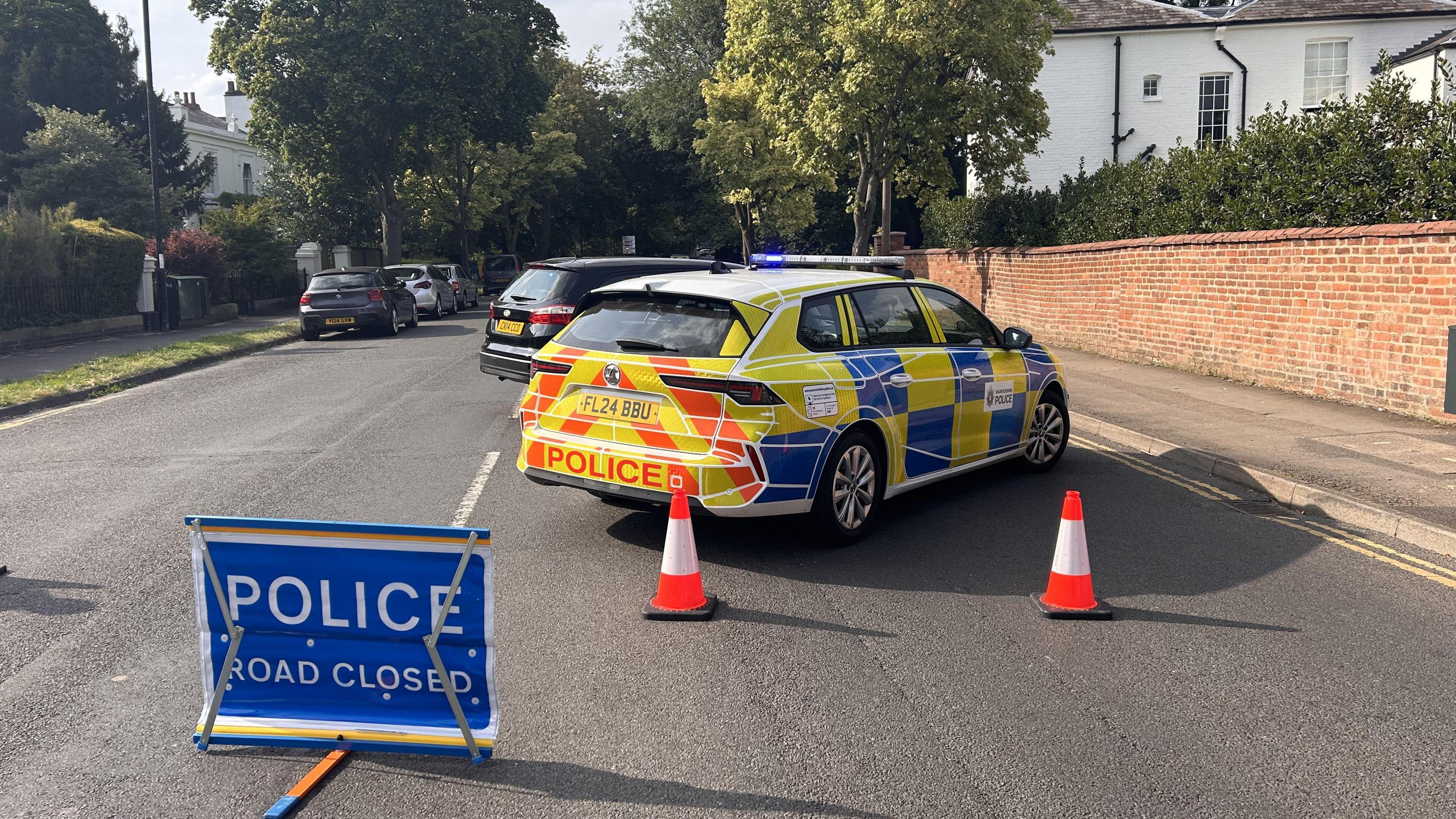 A police car cordoning off the road with orange cones and a blue sign that says 'police road closed'