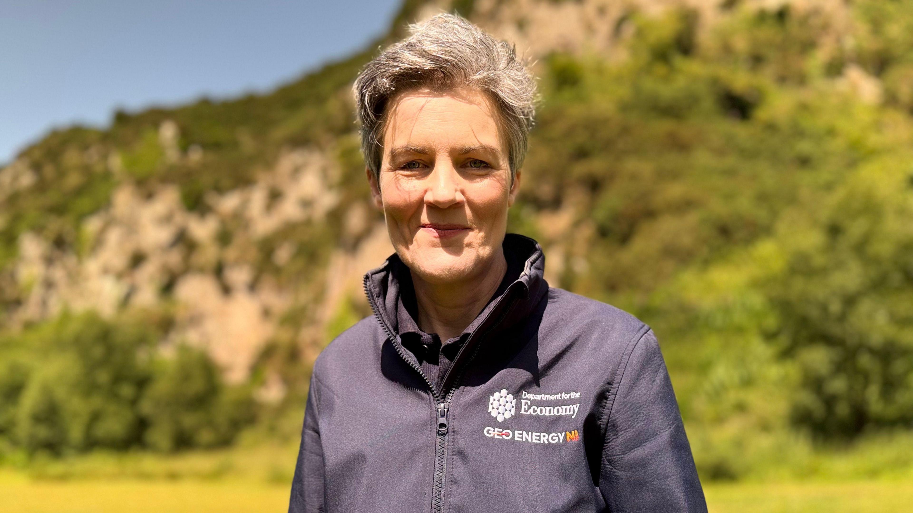 Dr Marie Cowan wears a navy jacket with a Department for the Economy logo on it. She is standing outside in front of a rock face and has short grey hair.