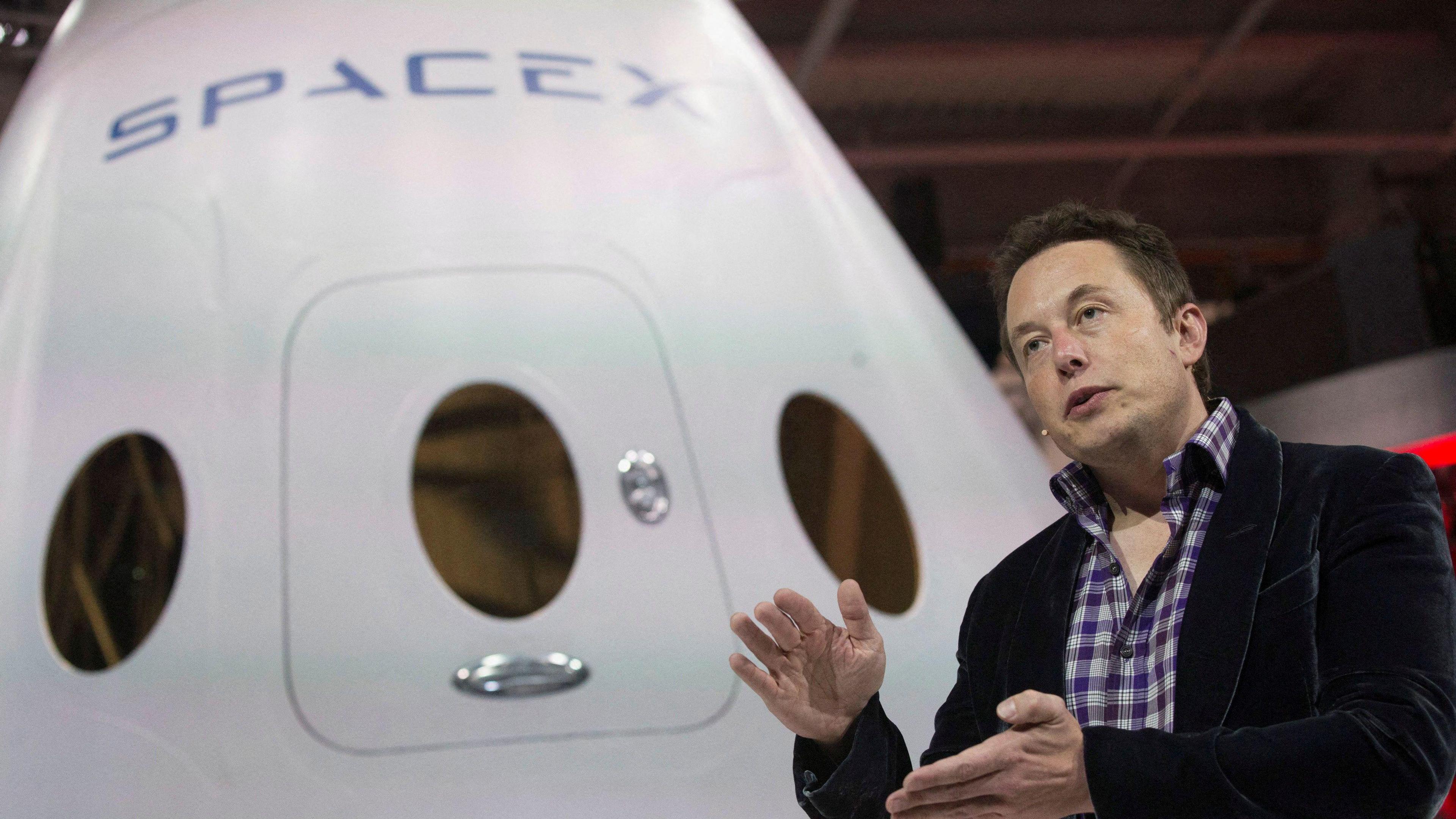 Elon Musk, who is wearing a blazer and a headset microphone, is speaking in front of a white spacecraft with the SpaceX branding on it.