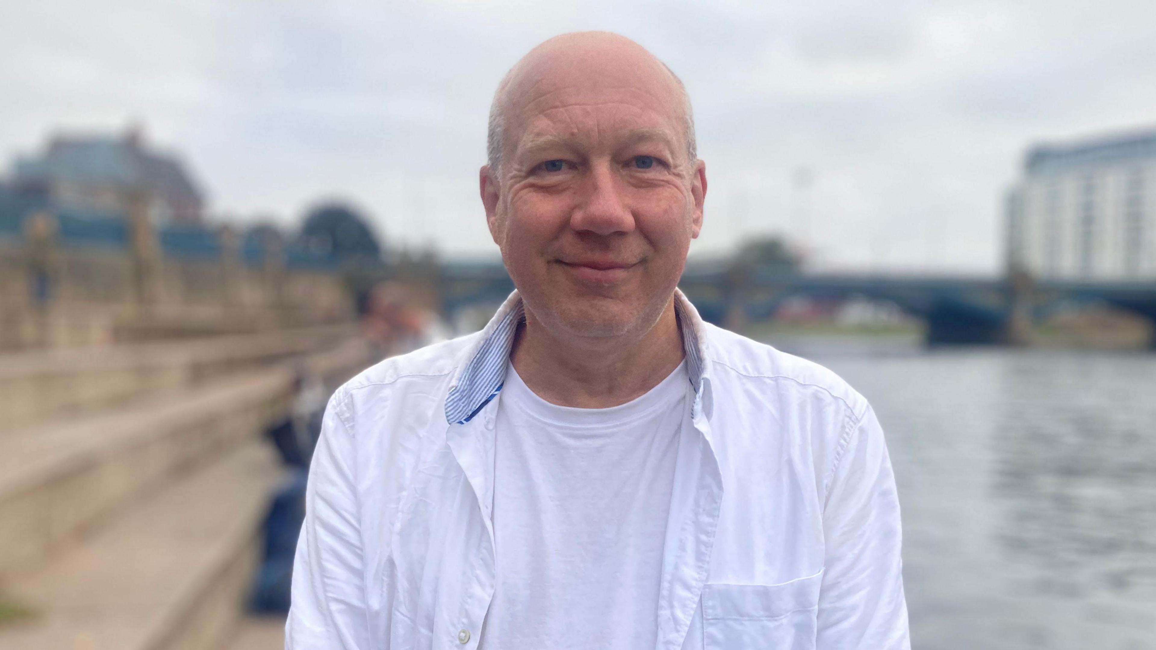 Simon Withers stands in front of the River Trent smiling for the picture. He is wearing a white shirt and white T-shirt underneath. His top half is pictured.