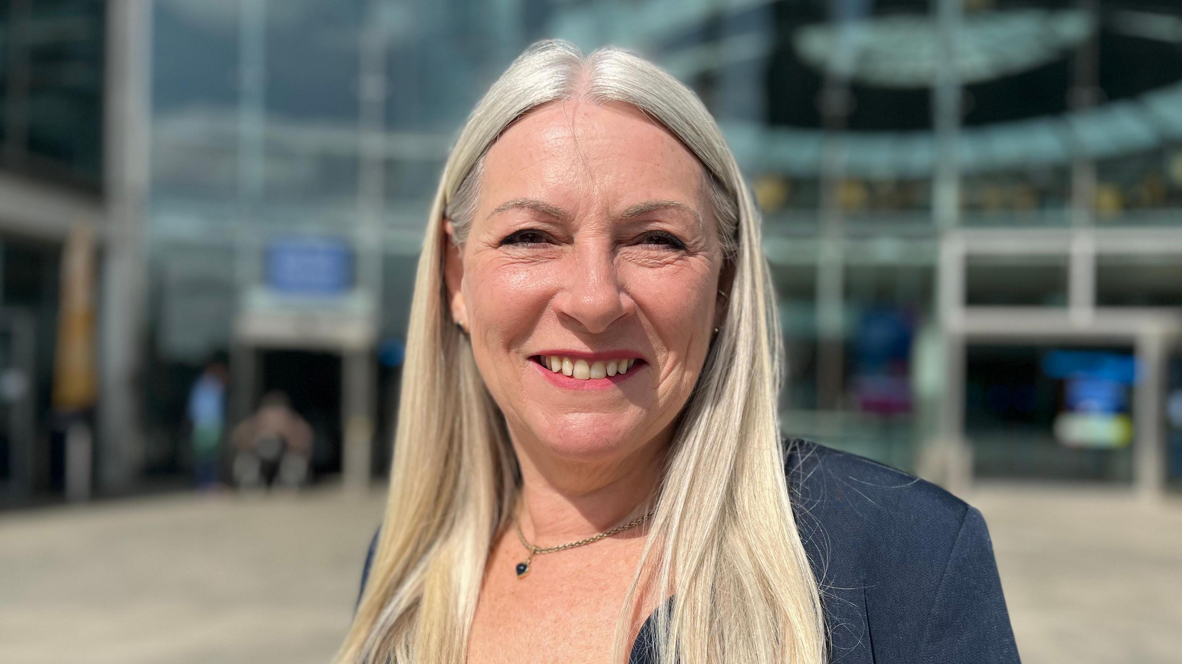 Kay Mason Billig smiling and wearing a dark blue suit jacket. It is clearly sunny weather. She is standing in front of the Norwich Forum building.