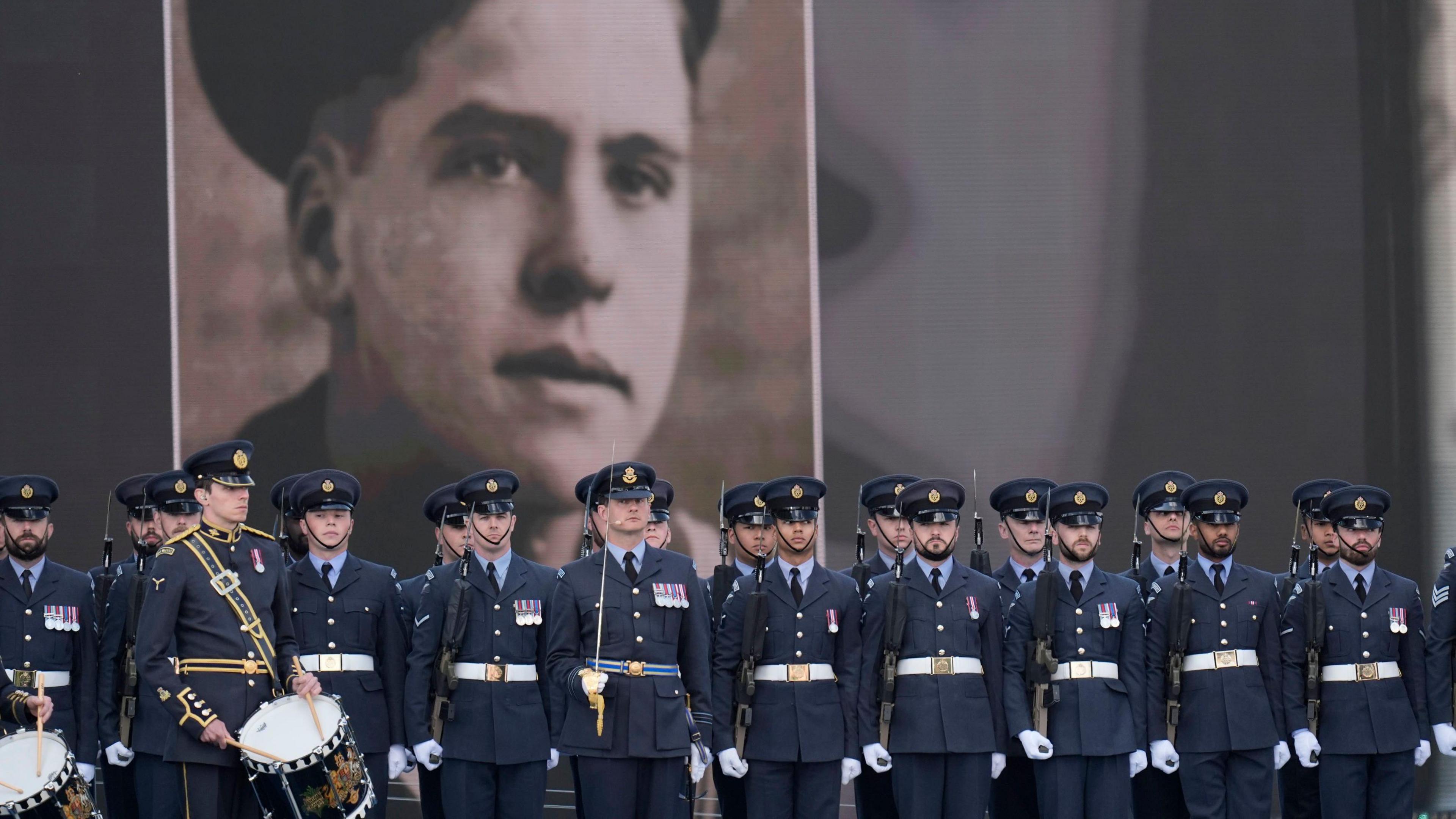 Members of the military on stage with guns in hand and some with drums stood in front of an image of a veteran