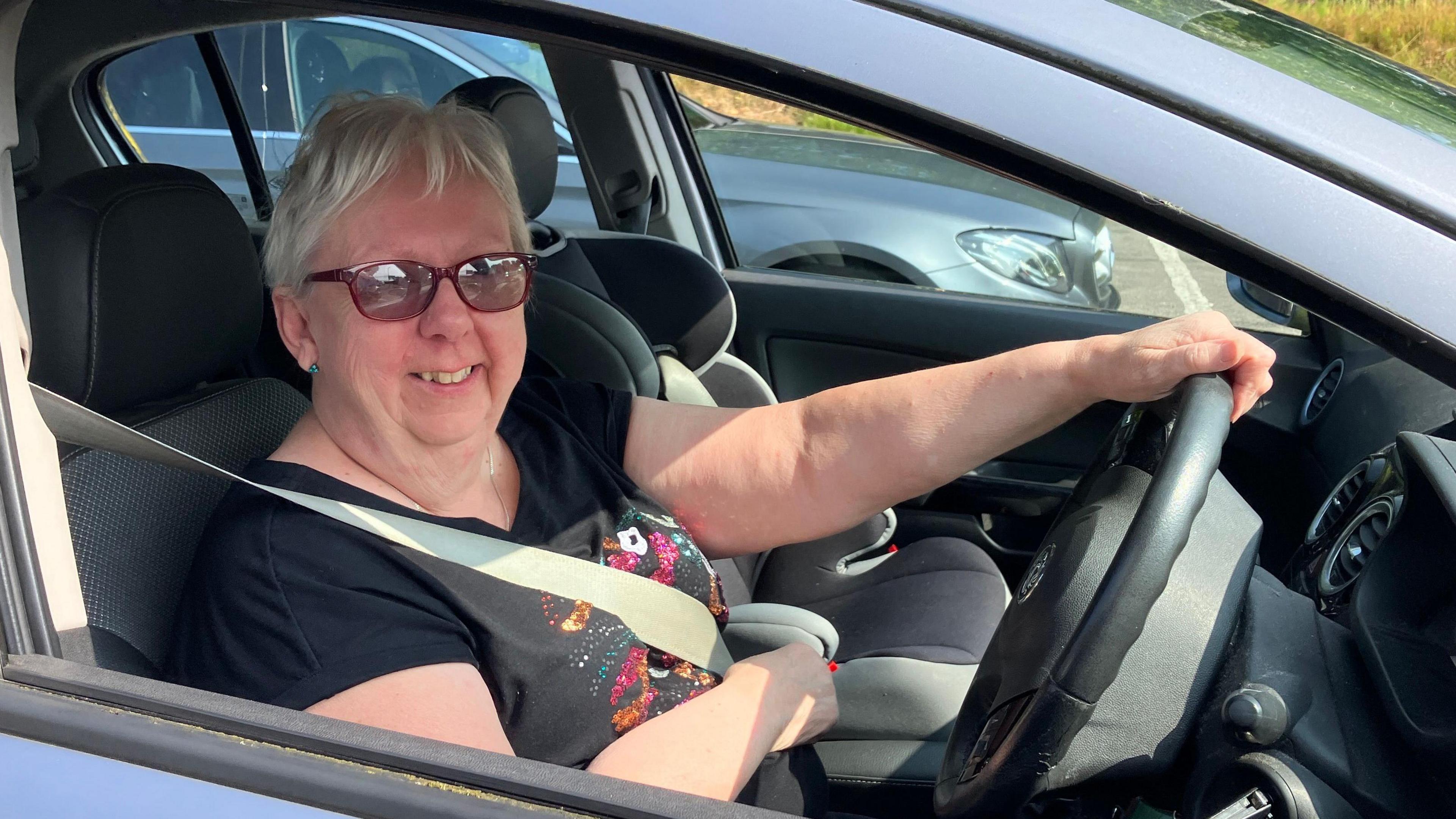 Fiona Lettsome in the driver seat of her car. She is smiling out the driver window and has short white hair. She is wearing sunglasses and a black t-shirt.