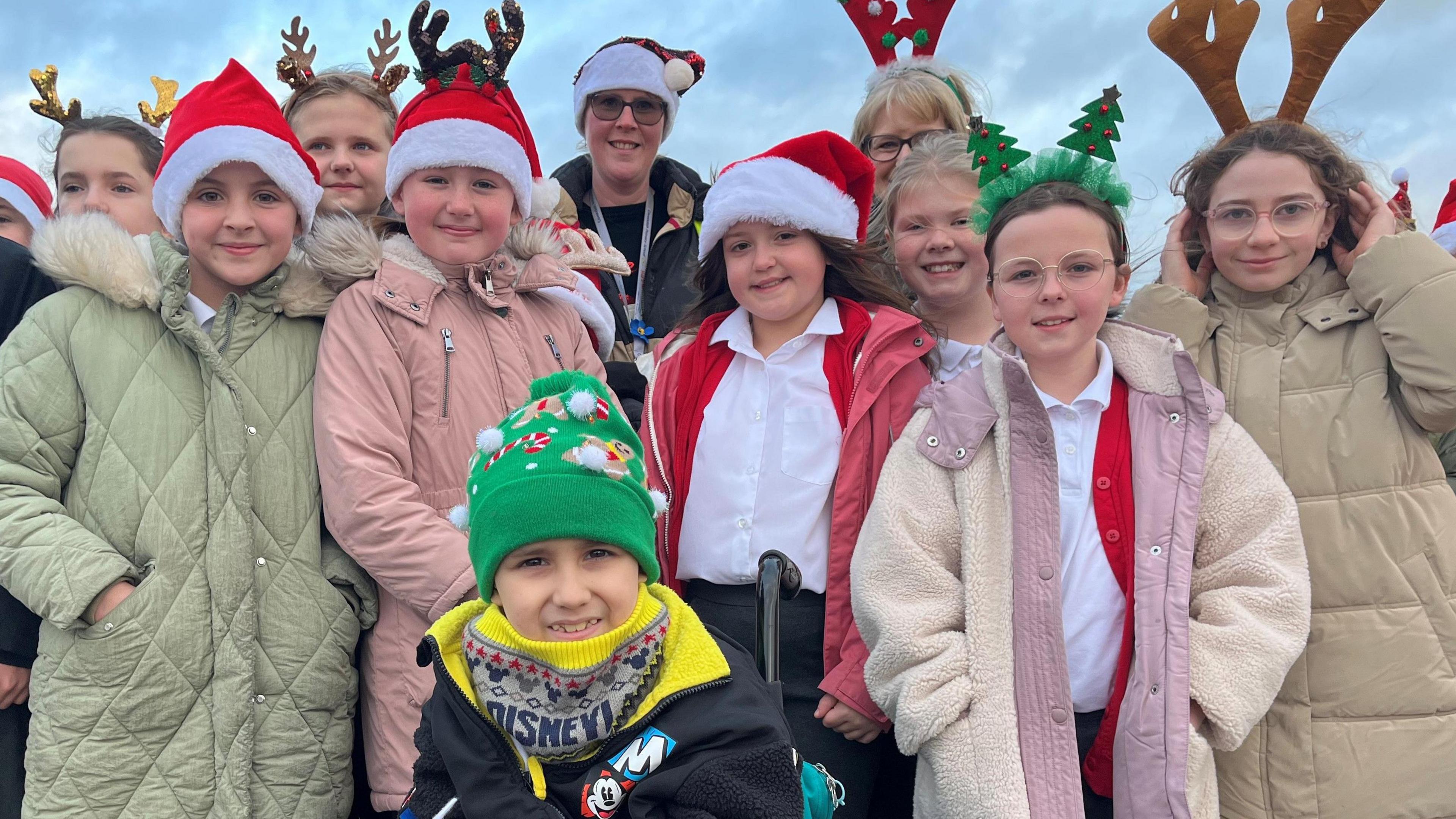 Isaac and nine of his classmates. Isaac is in the centre of the picture, wearing a black and yellow jacket and a green Christmas-themed bobble hat. Three of his classmates are wearing santa hats. Two school staff members are also at the back of the group. 