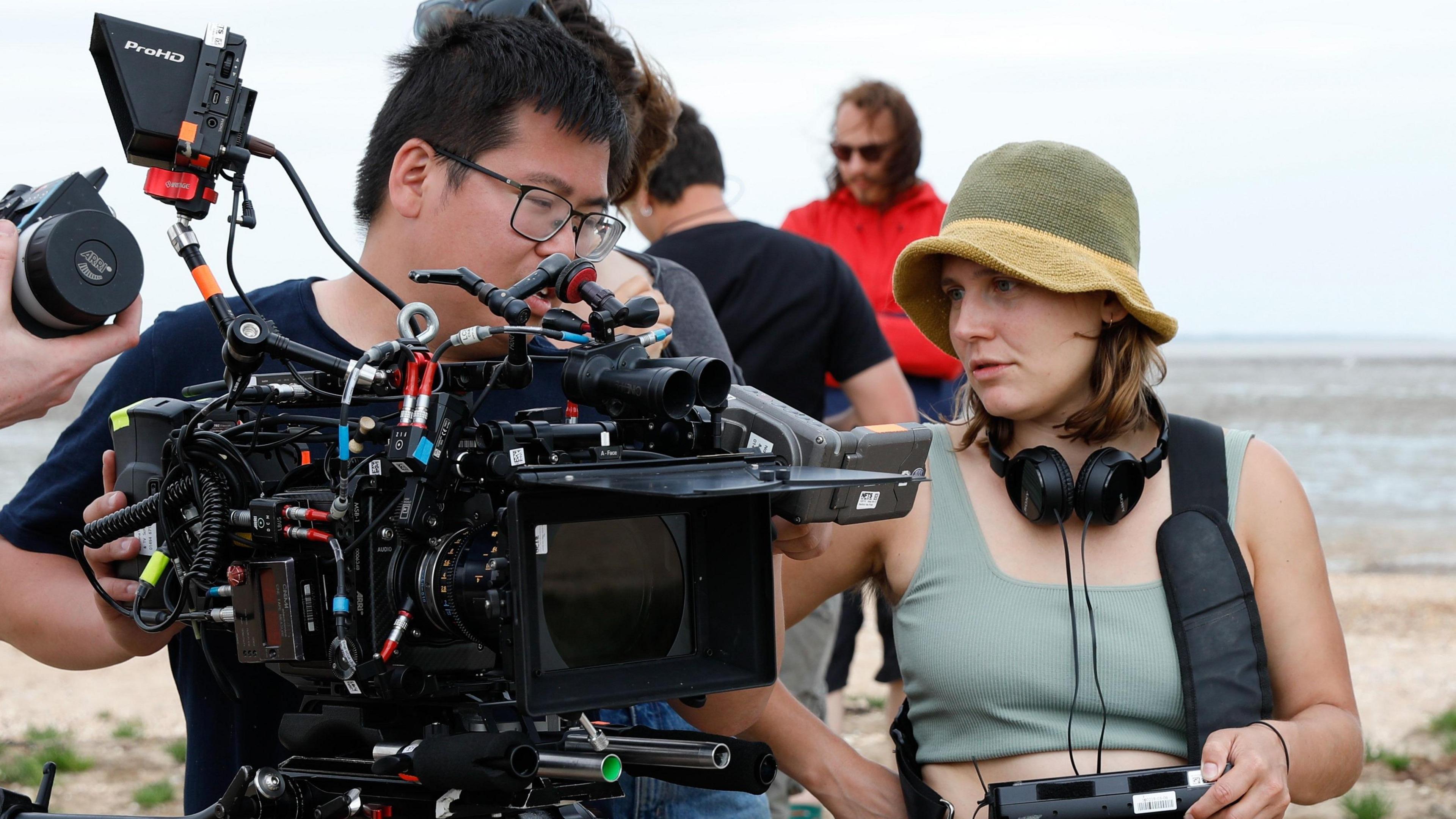 A man operates a camera to the left of the image as Hannah Renton looks at it. Behind them is a beach and the sea.