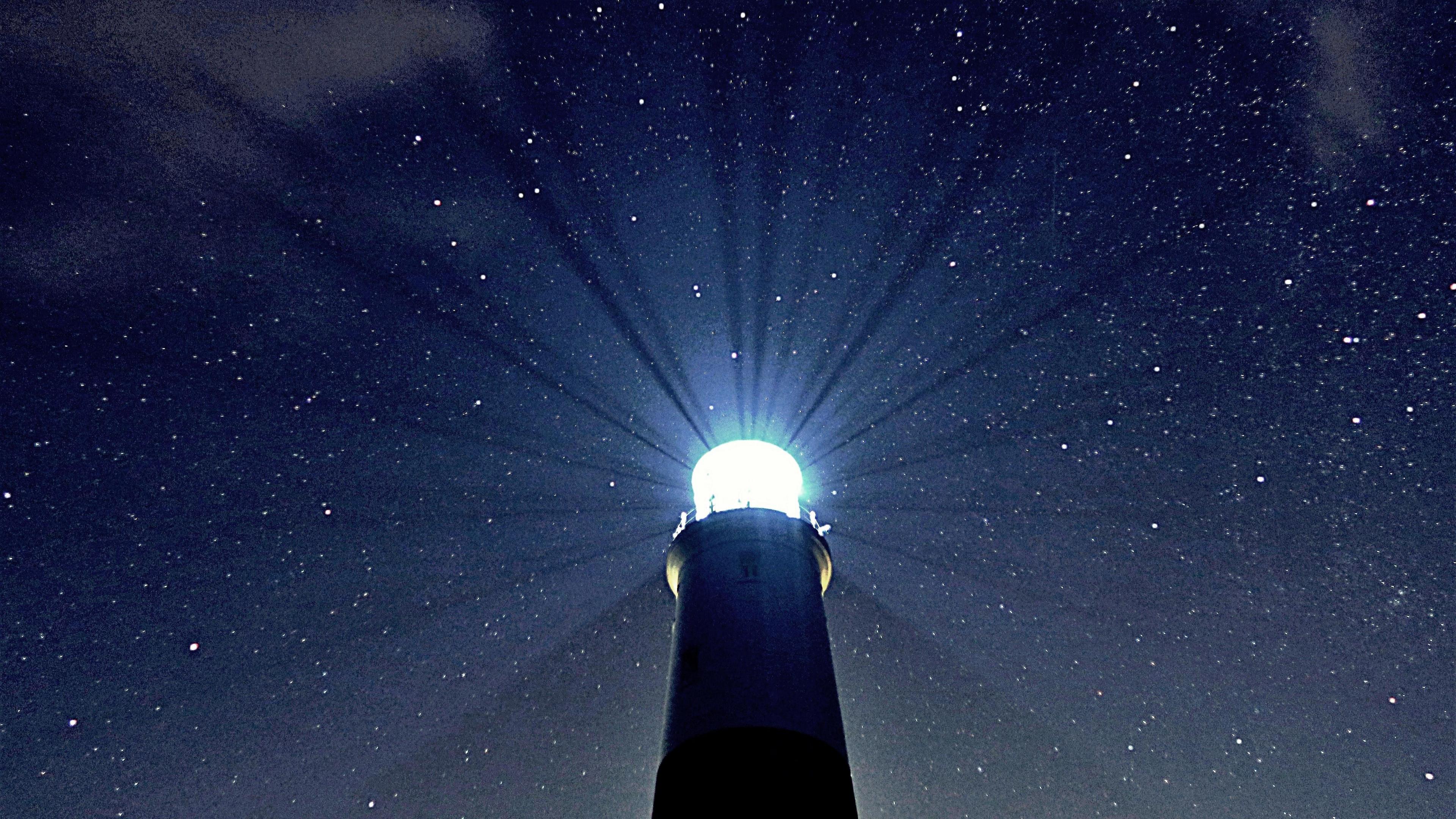 The sky is pitch black but studded with stars. In the centre stands the top of a lighthouse with its light shining bright. You can even see beams of light being cast from the lighthouse into the night sky.