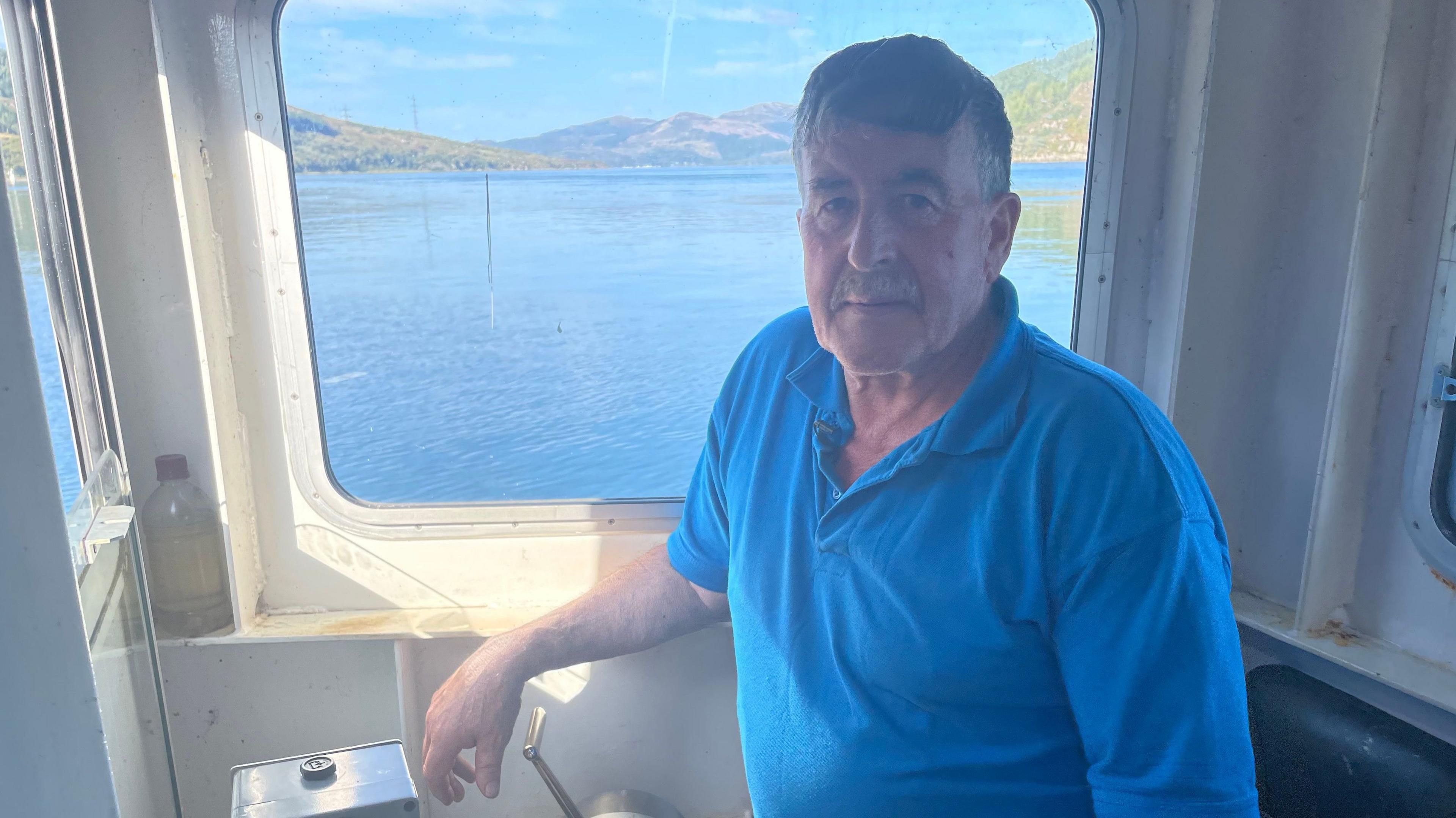 Fear le falt goirid liath agus stais liath ann an taigh-cuibhle bàta.  Tha lèine ghorm air.  Chi thu am muir tron uinneig air a chùlaibh agus tìr le craobhan.  