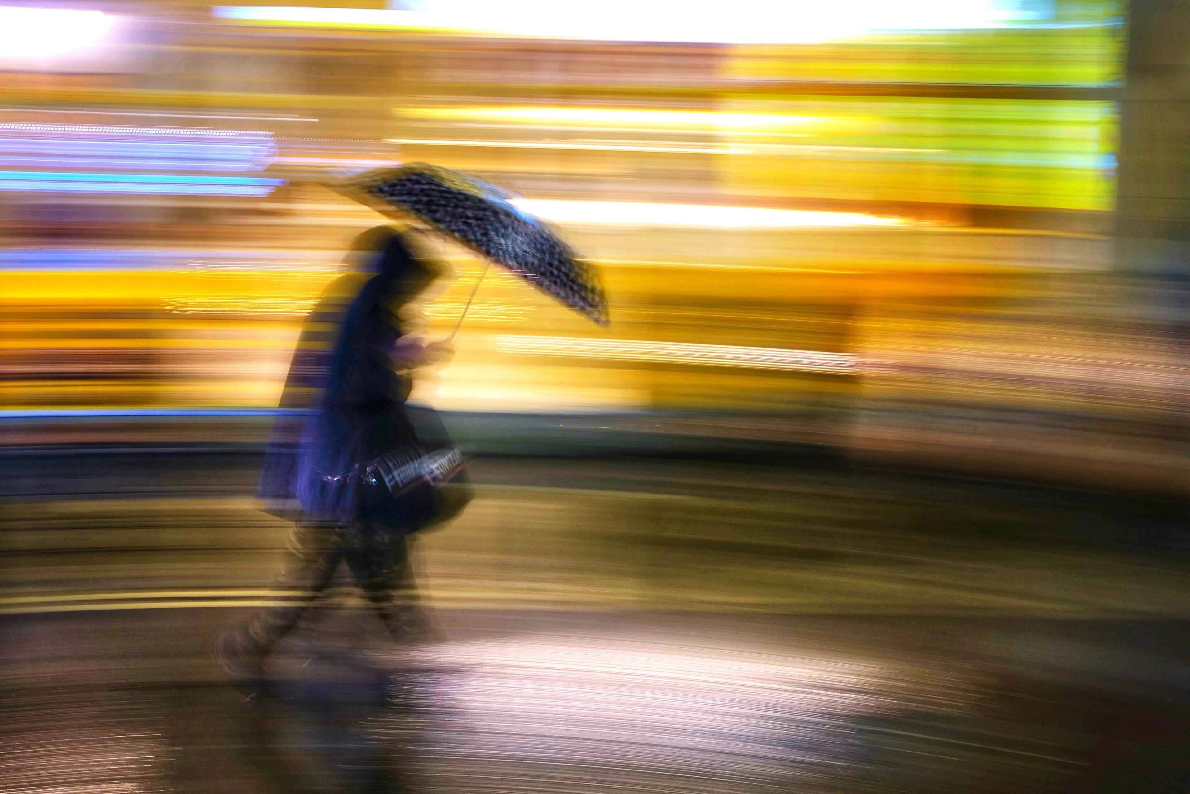 A person with an umbrella passes bright lights. The person has their head down and is walking against the rain.