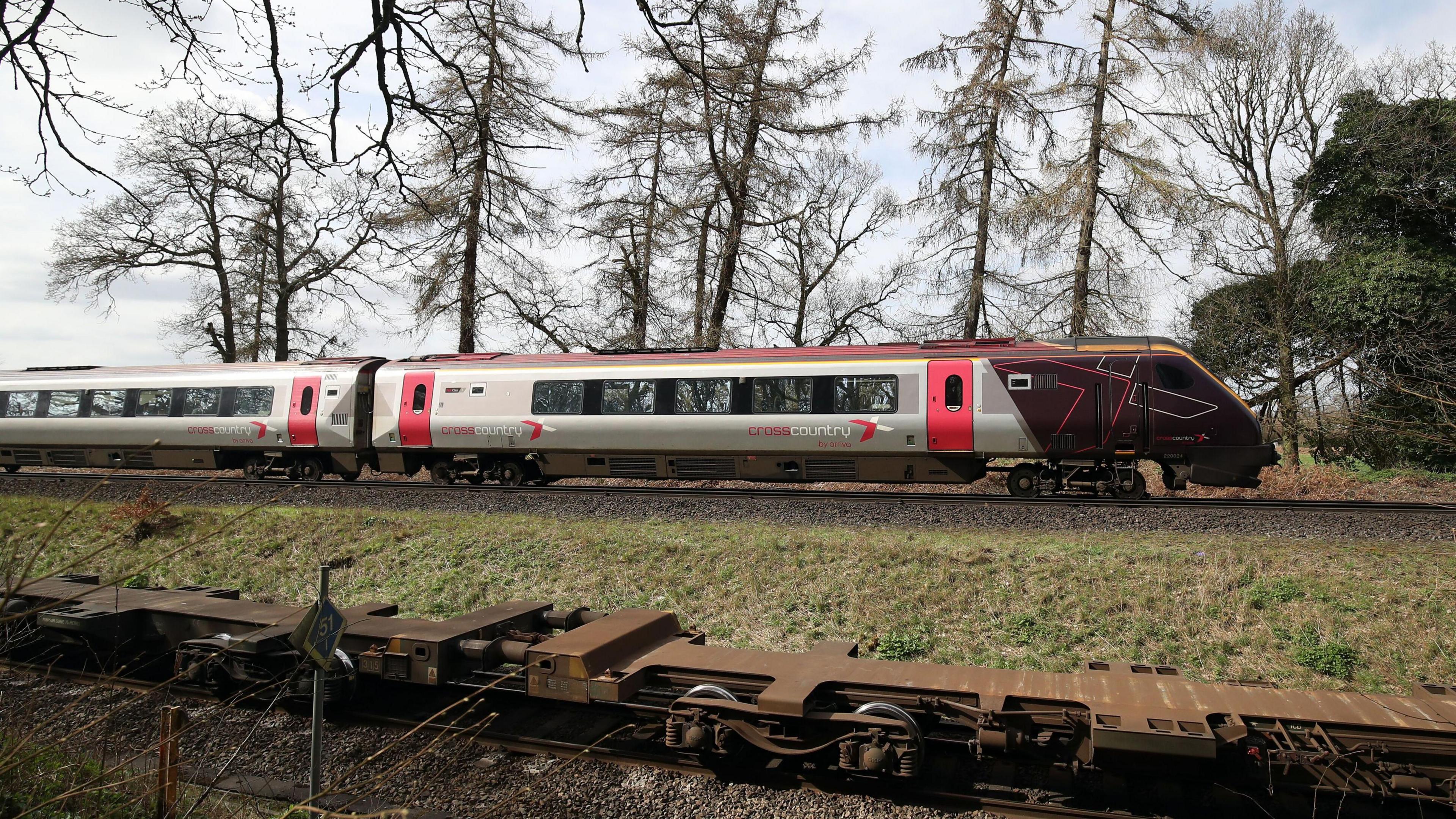 The cab and first carriage of a CrossCountry train travels on tracks in the countryside. In the background trees and fields can be seen.