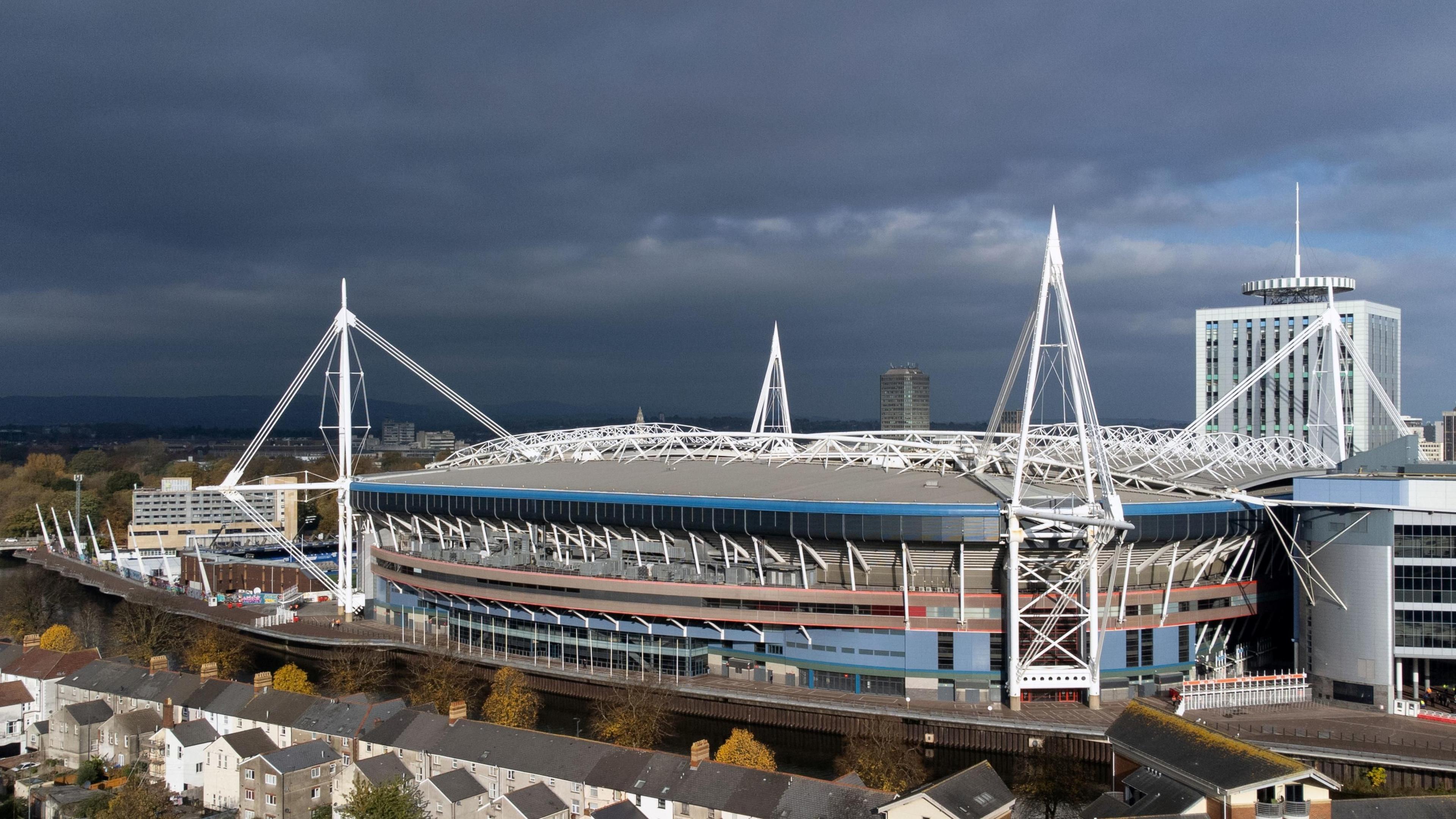 An aerial view of the Principality Stadium. Stormy grey skies are seen behind it, while in front runs the river and trees and a row of terraced houses.
