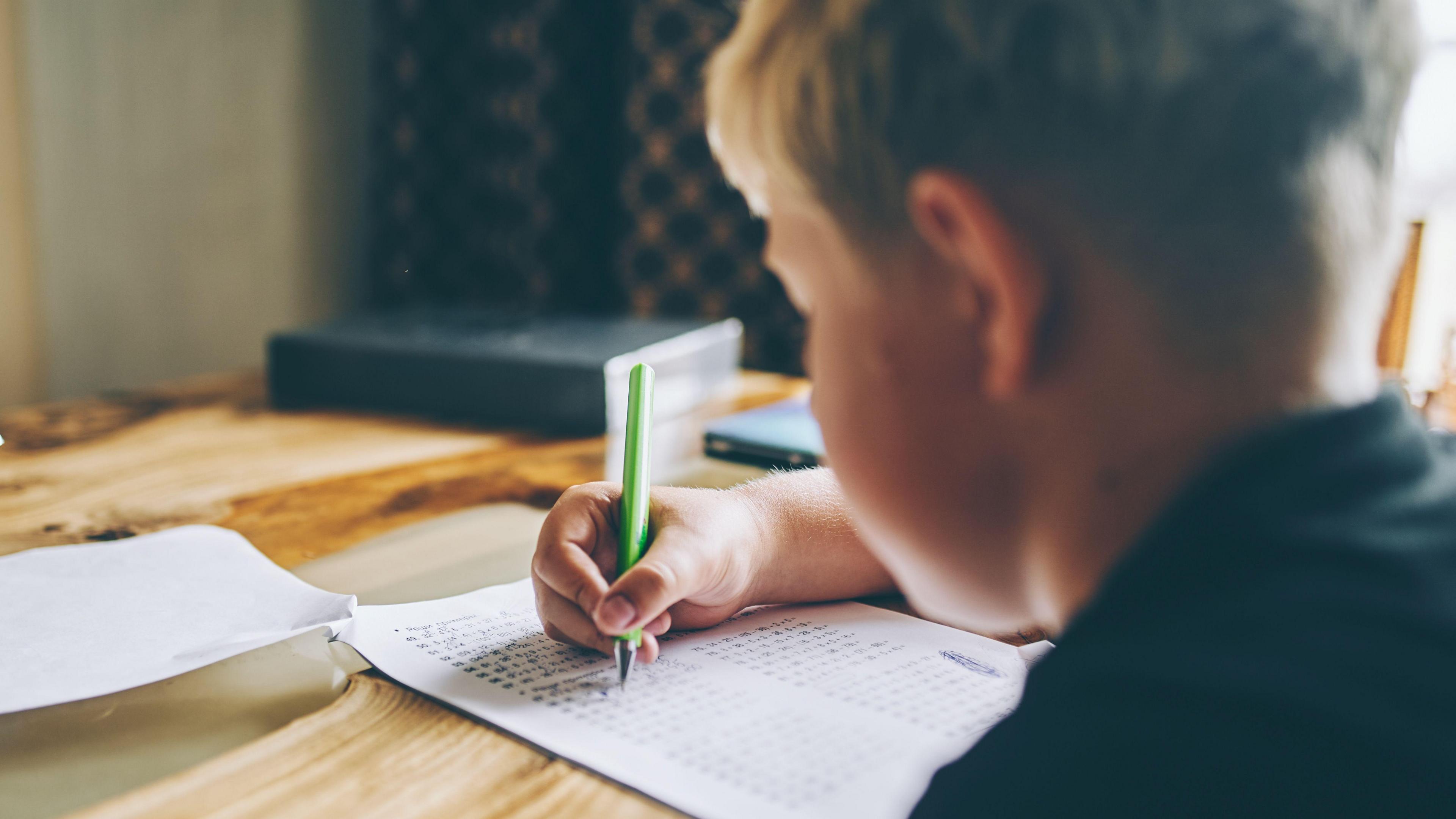 A boy doing school work at a kitchen table. He's holding a green pen. The camera is behind him. He has fair hair and is wearing a black top.