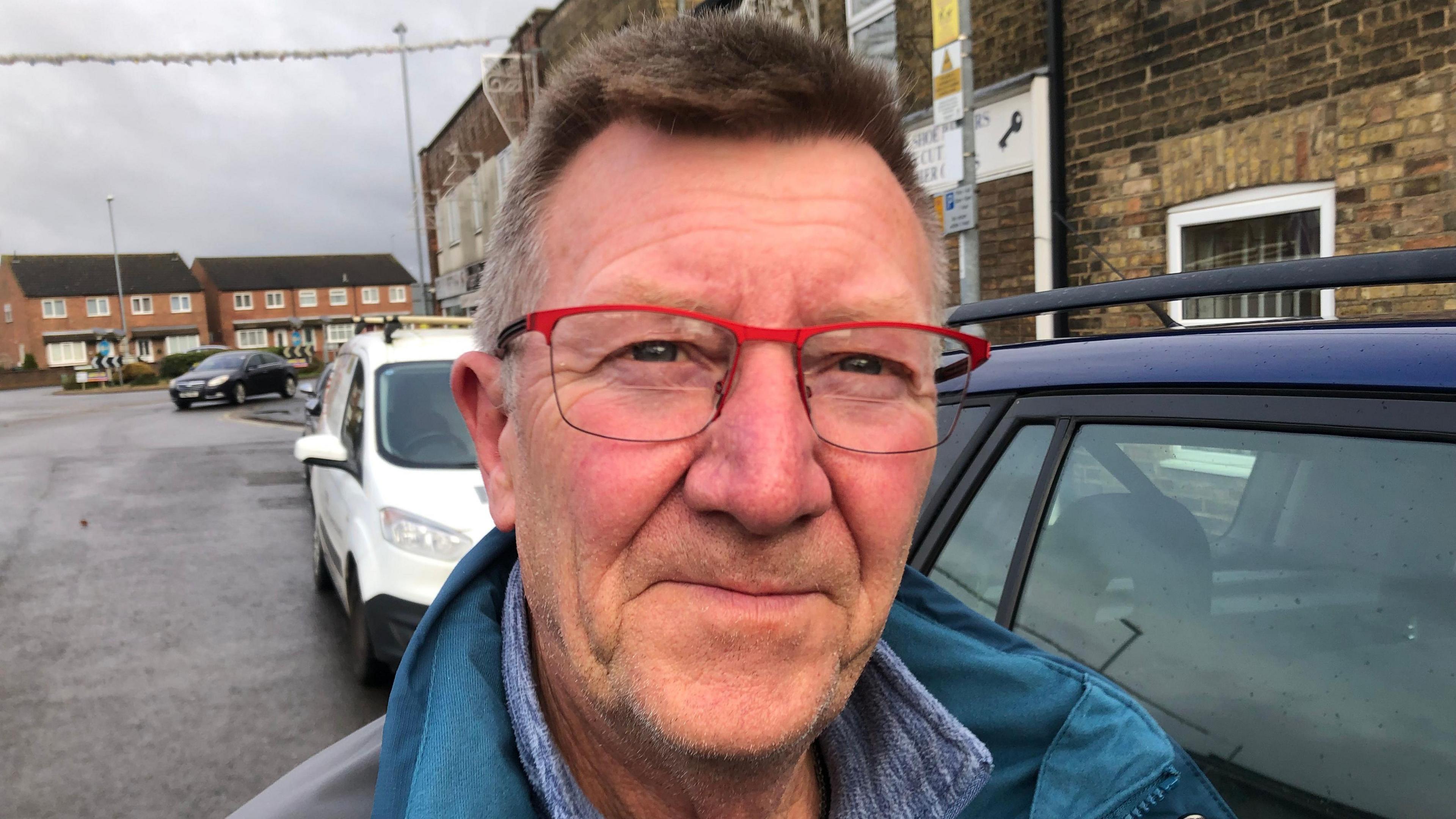 Phil Slinger smiles at the camera. He is wearing a blue coat and glasses with a red frame. He is standing in a street with a number of cars parked alongside the pavement behind him. 