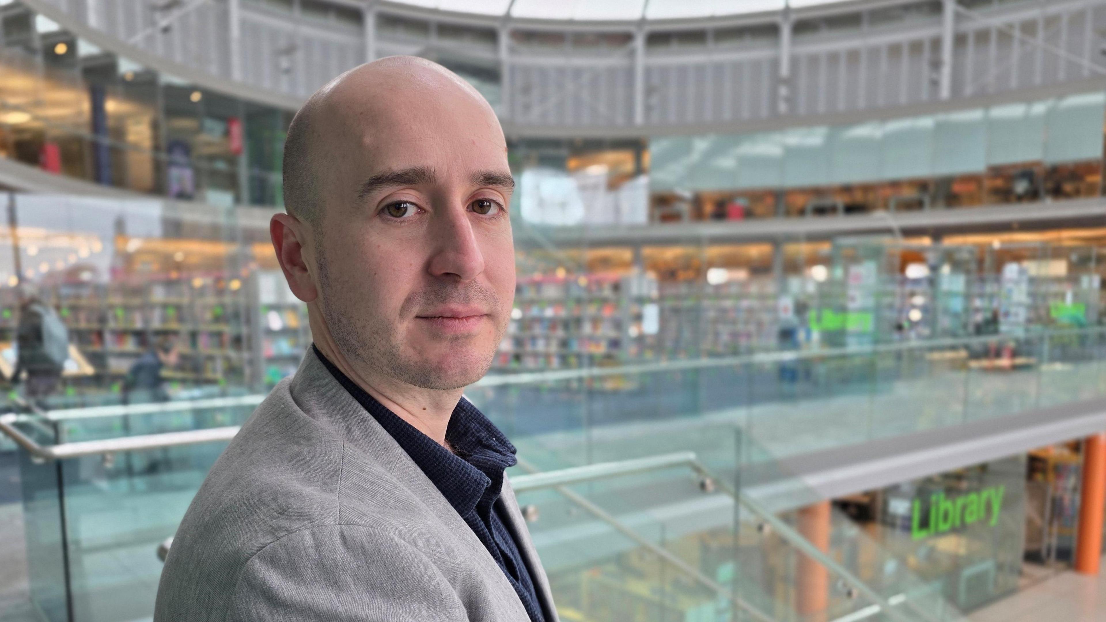 Ben Goldsborough is looking at the camera over his right shoulder. He has a grey suit jacket on over a blue shirt. He is standing on a glass internal balcony, with the library visible behind him.