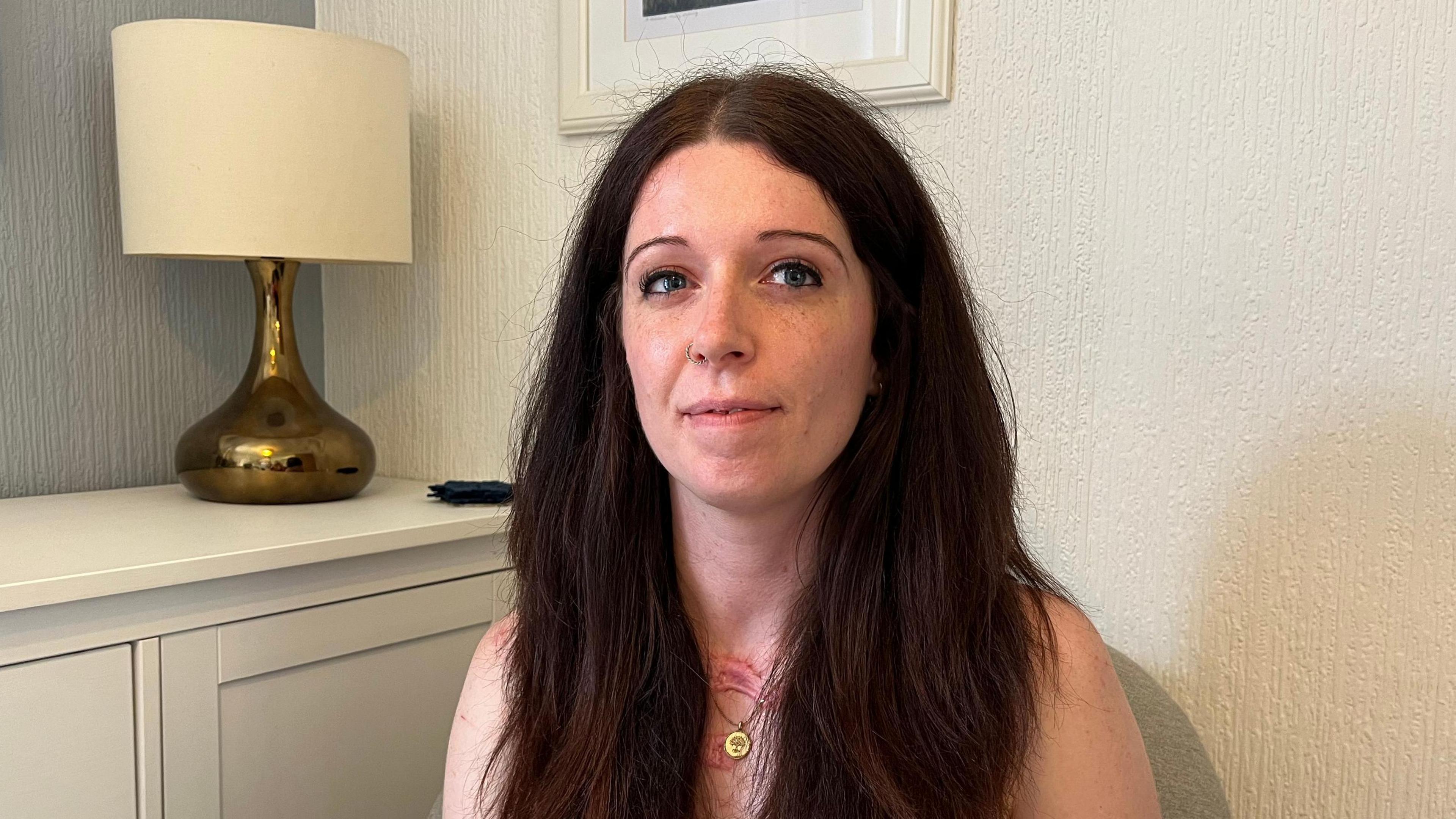 A woman with long brown hair smiles at the camera. She has two red scars on her chest. She sits in a living room with white walls and a white lamp on a table behind her.