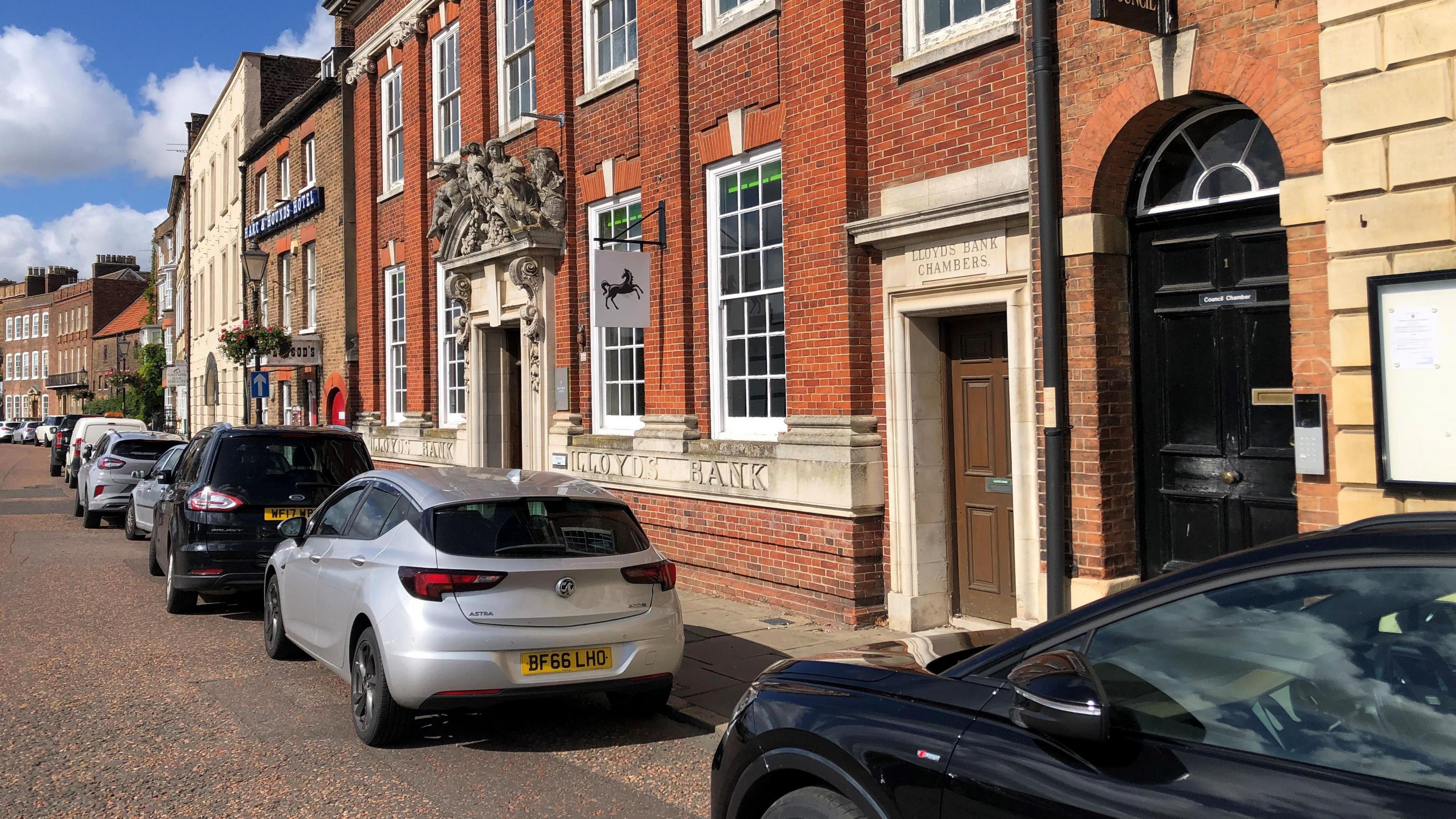 The outside of Lloyds Bank branch in Wisbech, a red-brick building with a row of cars parked outside. 