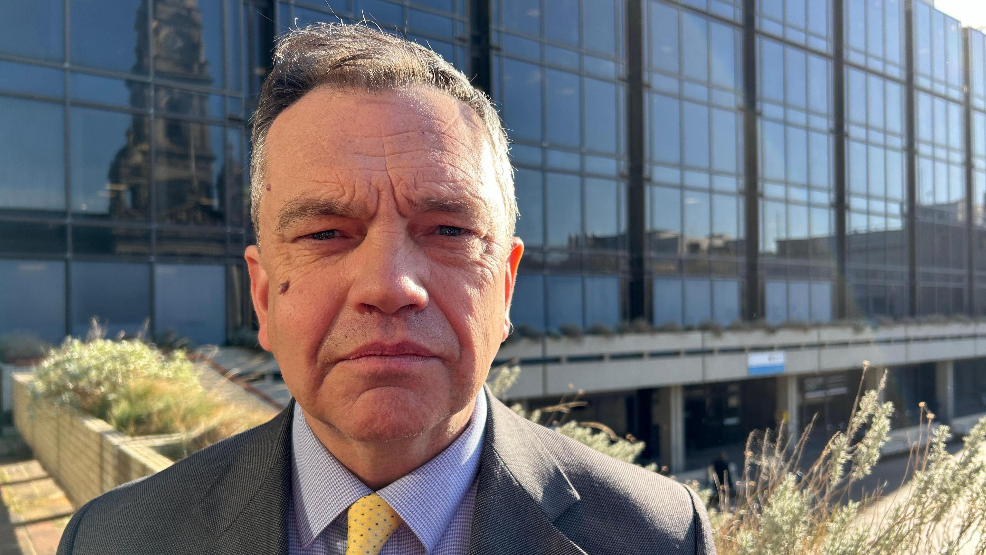 Councillor Darren Sanders outside the council building. He has grey hair, and wears a grey suit with a blue shirt and yellow tie. 