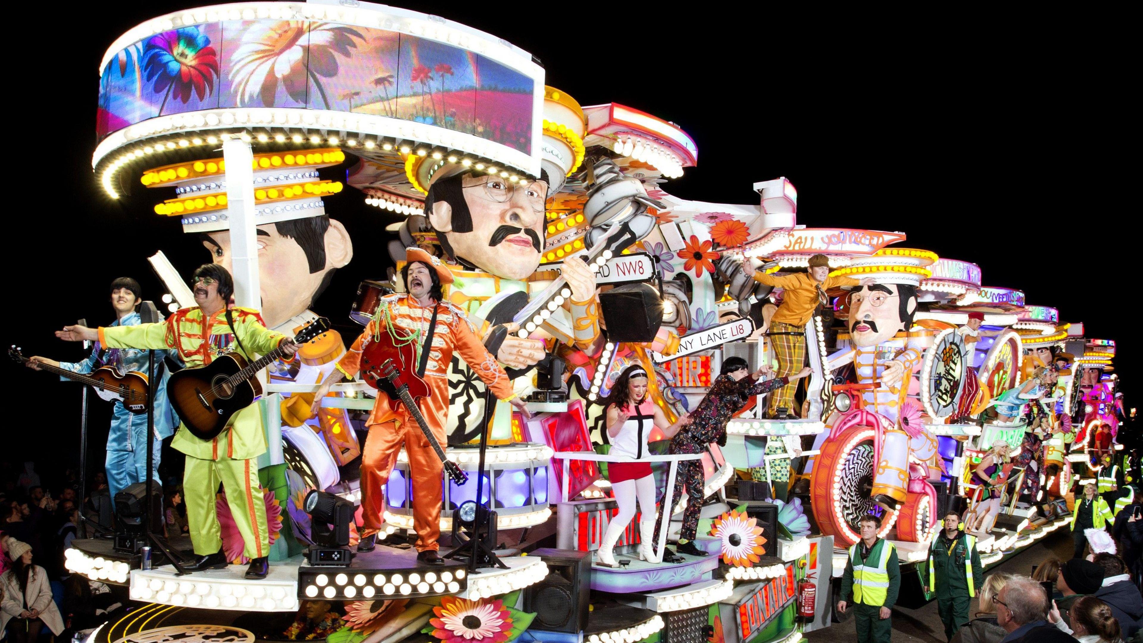 An illuminated carnival cart with many colours and people standing on it holding guitars.