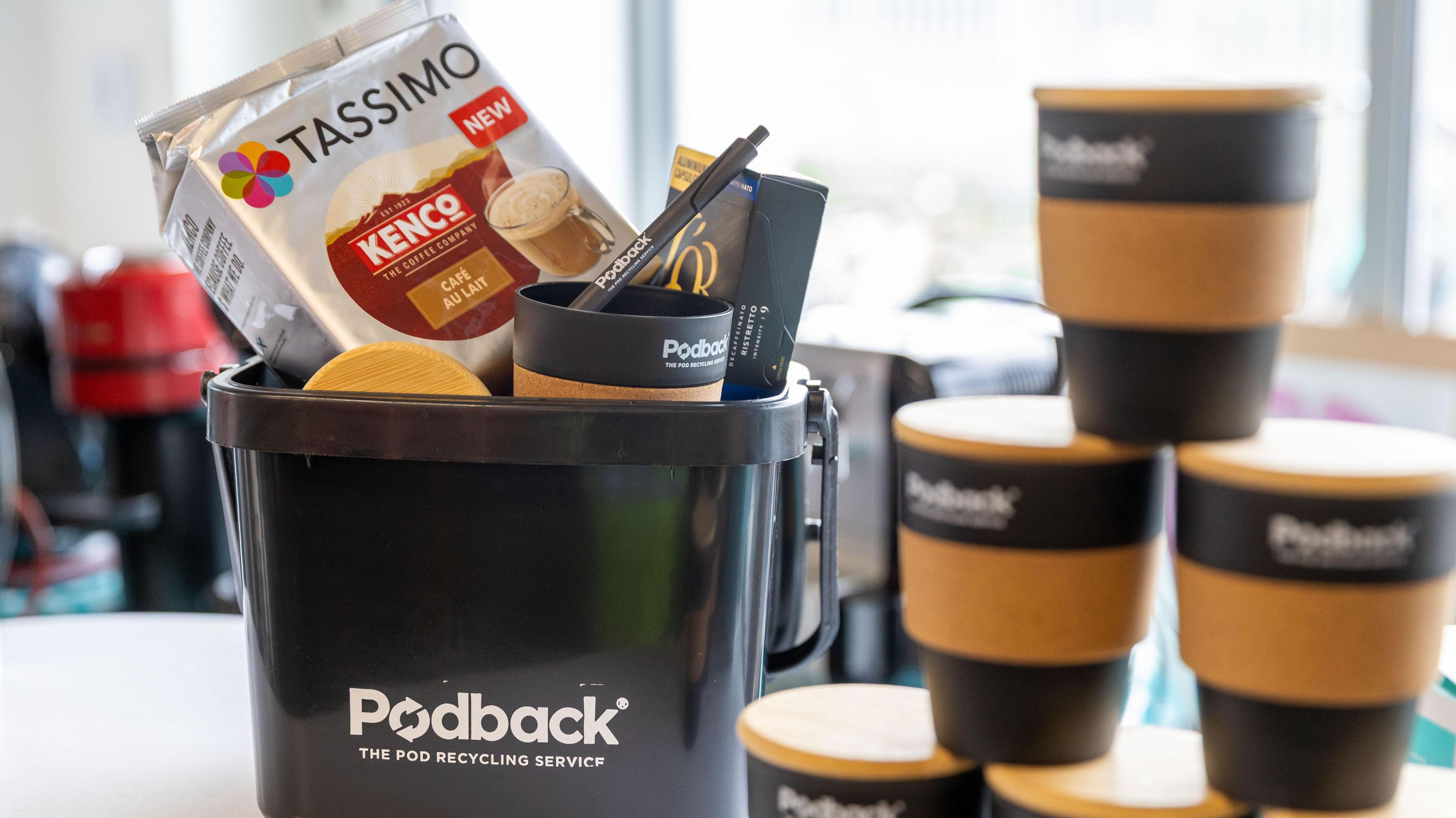 A black Podback container with coffee making paraphernalia inside it and reusable coffee cups next to the container branded with "Podback"