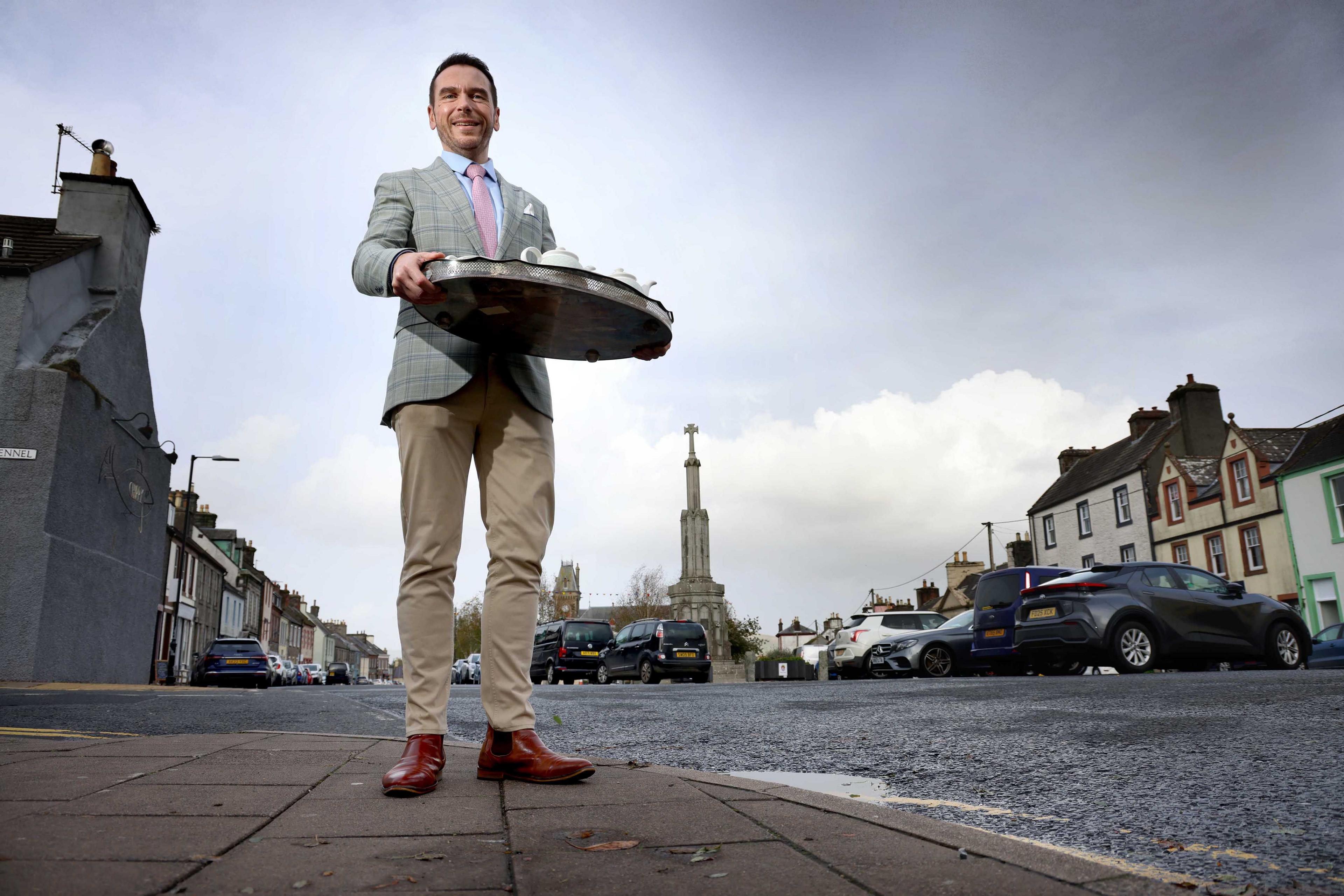 Grant Harrold in smart clothing carrying a tray with a tea service on it. He is standing on a pavement in a wide street and looking down at the camera.
