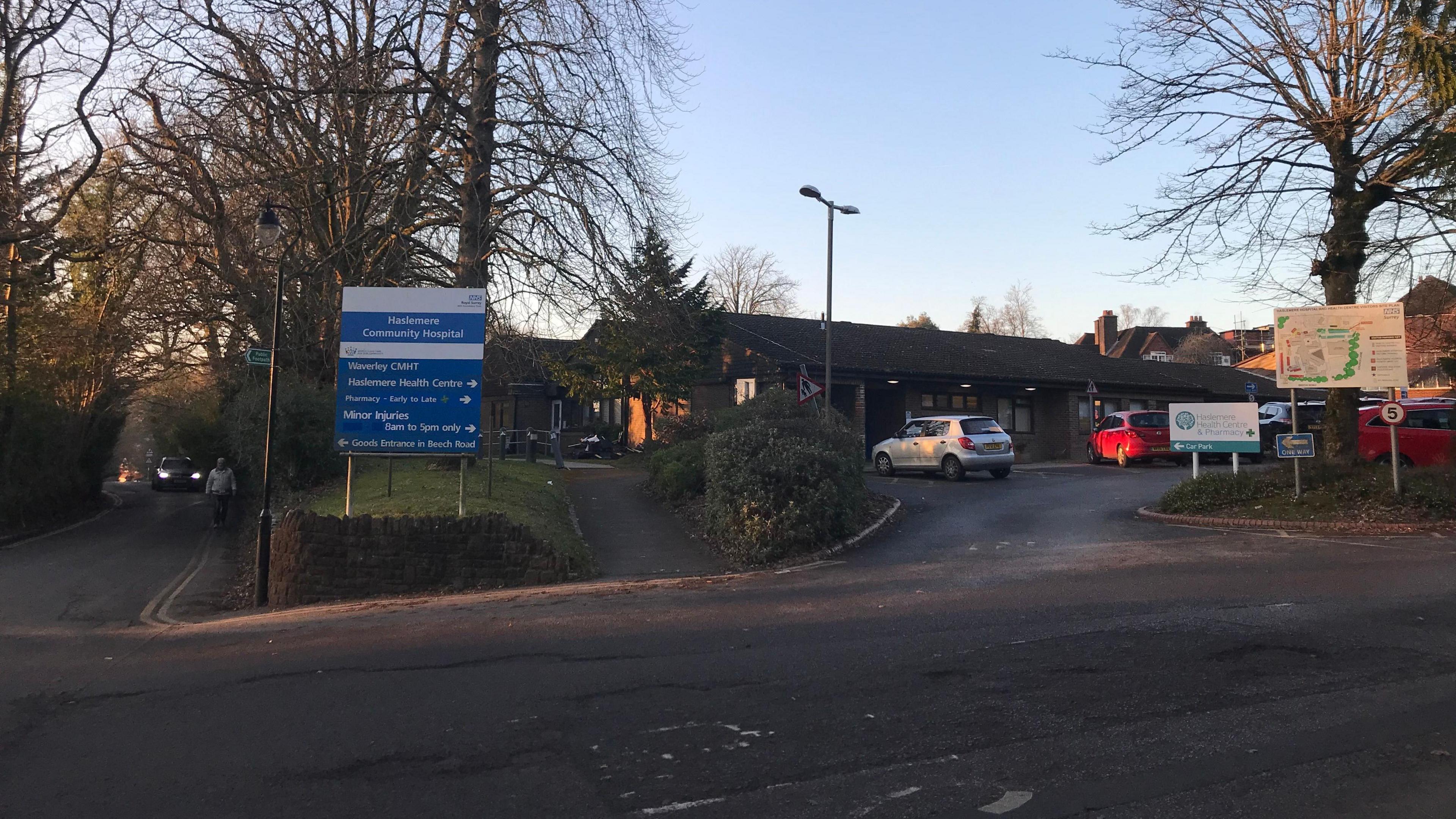 The exterior of Haslemere Community Hospital, showing the entrance sign, the driveway and two cars, one grey and one red, parked outside the building.