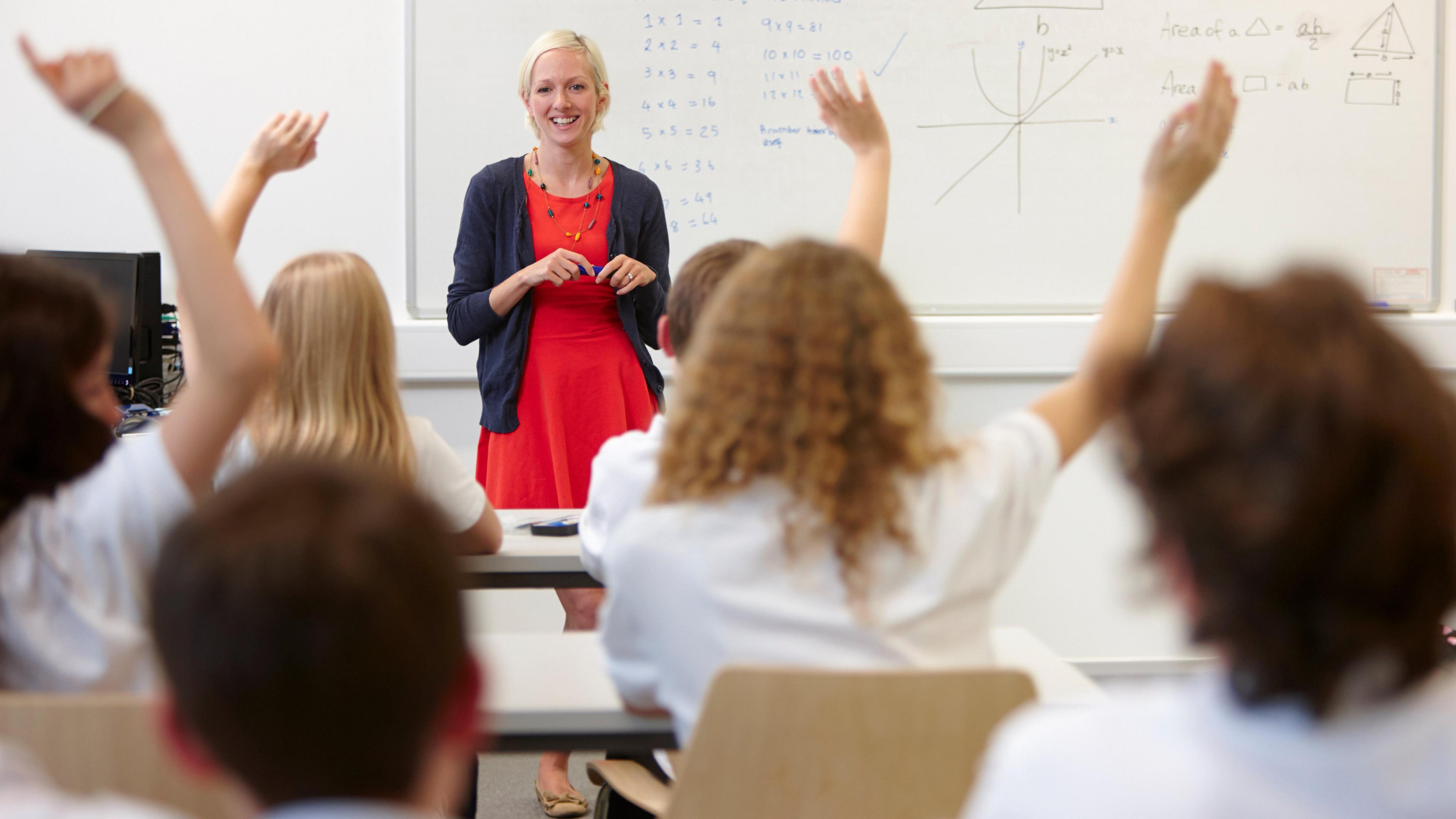 A classroom with children in school uniform with their backs to the camera facing their teacher who is smiling. The children all have their hands up and there is a maths equation on the board.