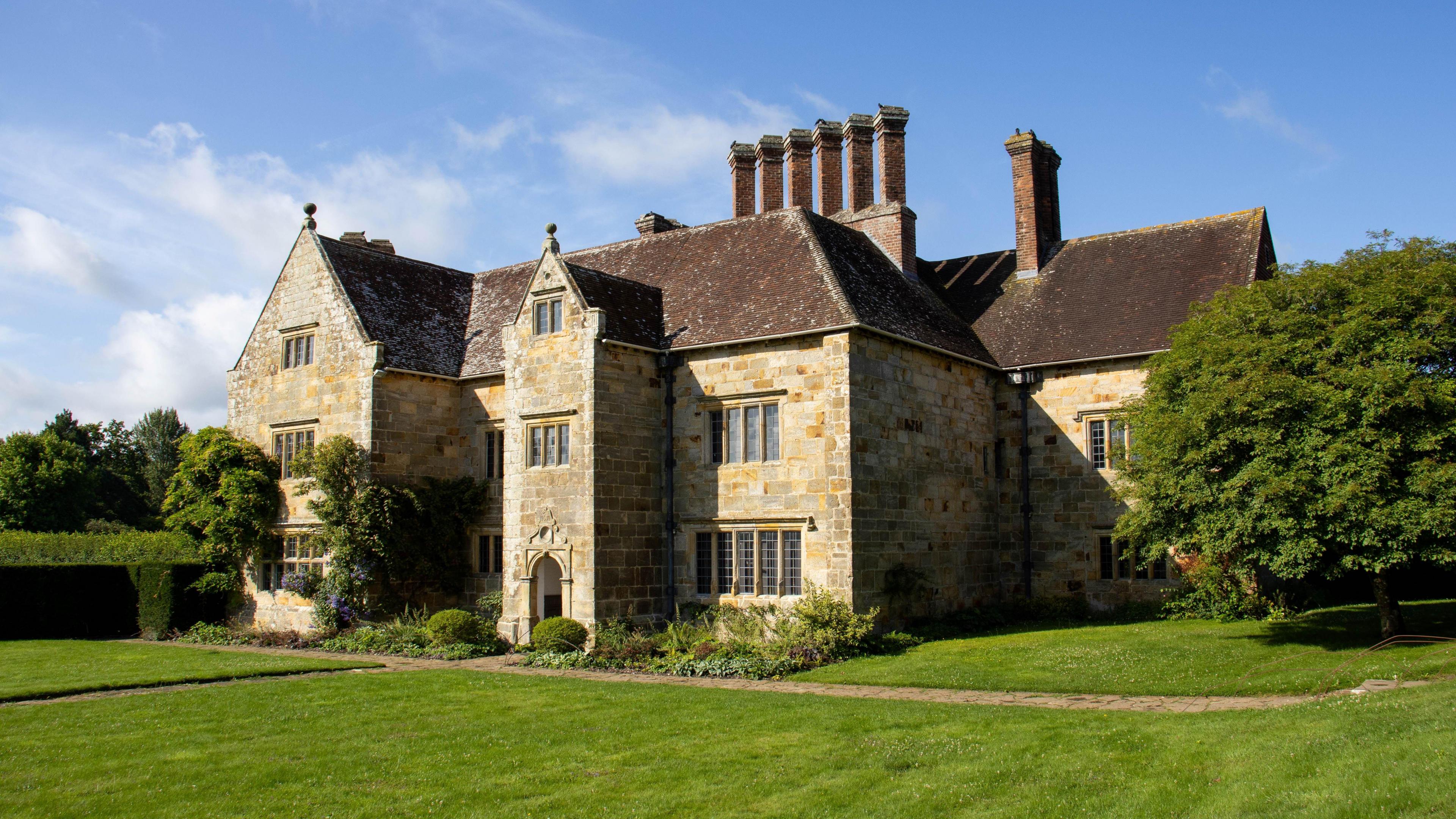 The front of the Bateman's house, it is sunny. The house is made of stone and has a brown tiled roof, there are seven chimneys. 