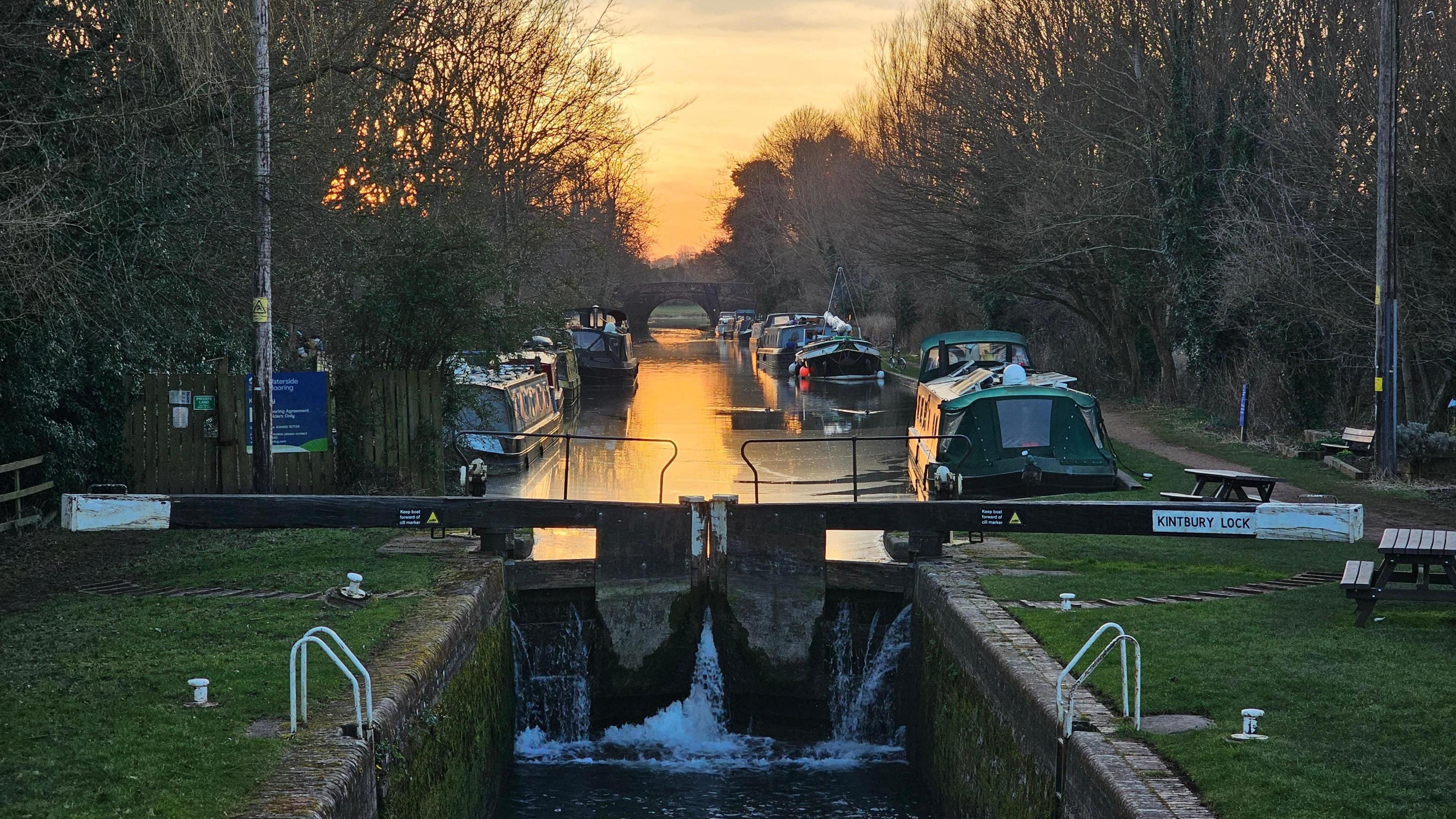 The sunsets over the canal. In the foreground is a lock gate. Behind the glowing orange sky is reflected in the water. The canal has boats moored on either side. The bank has trees and grass next to the tow path. 