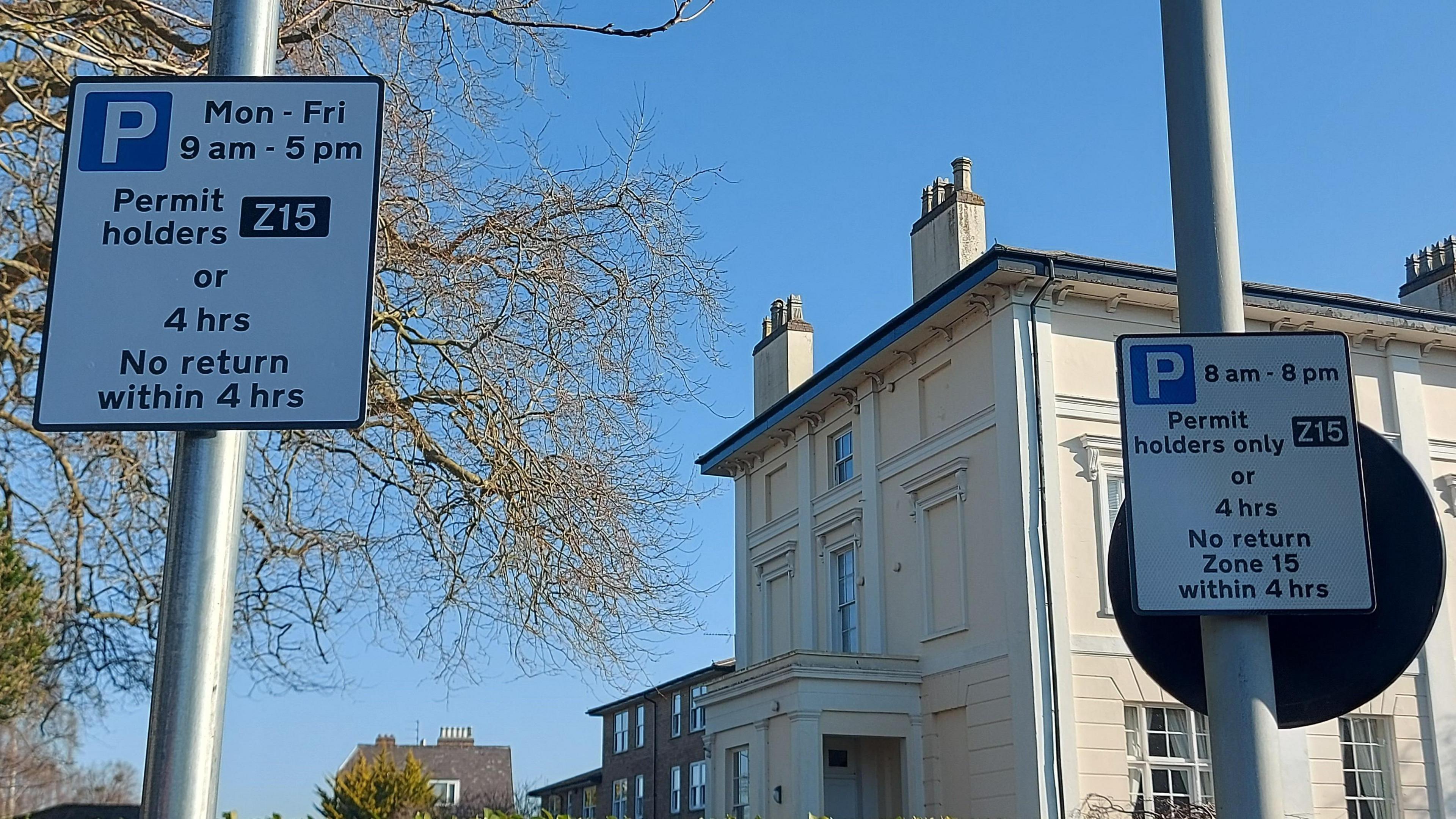 Two parking permit holder signs pictured against a bright blue sky on a winter day. There are bare trees in the background and a large cream-coloured building.